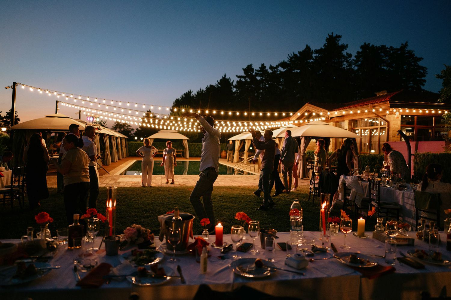String lights illuminate an outdoor venue at dusk with party tents and dancing guests.