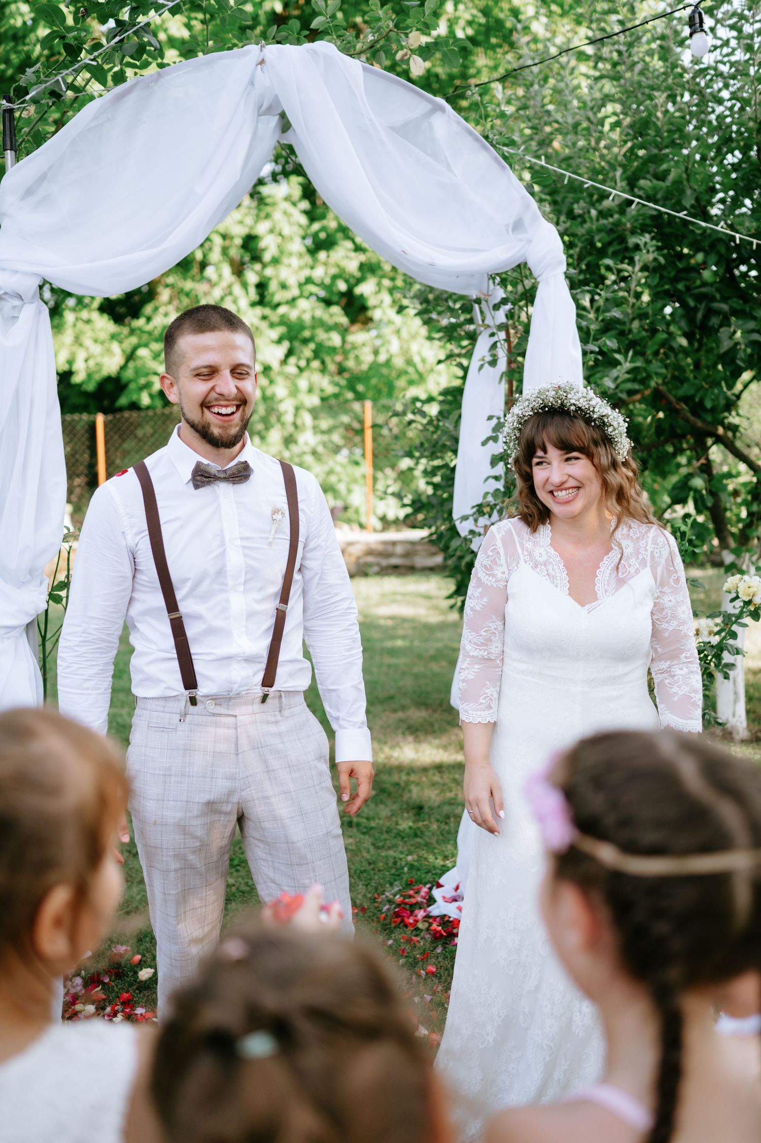 Wedding couple sharing a joyful moment under a white fabric arch surrounded by greenery.