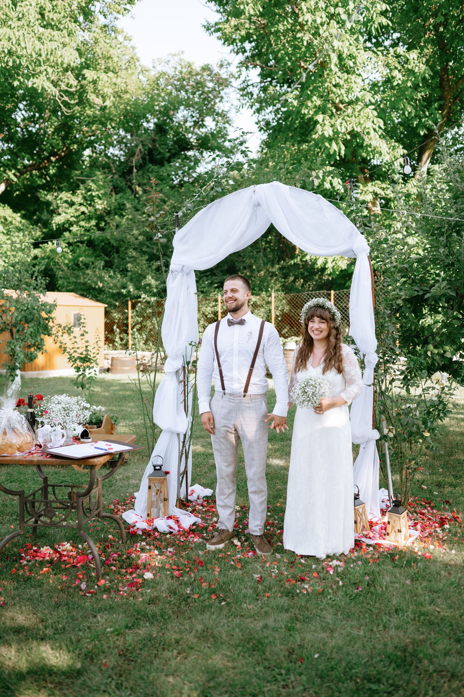 Elegant backyard wedding scene with white draped arch and rustic decor among lush green trees.