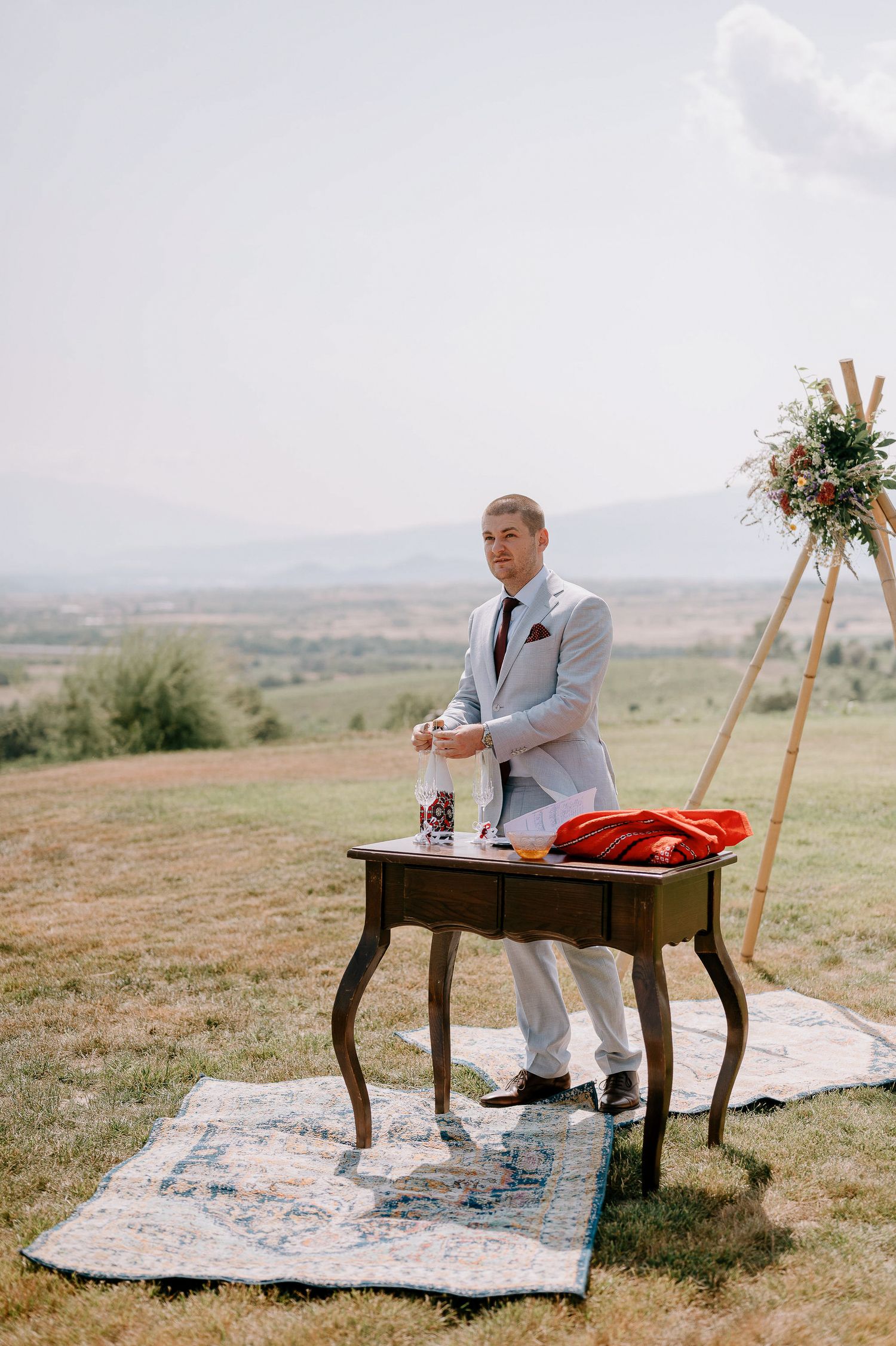 Person in light gray suit stands at antique table on hillside with floral arrangement.