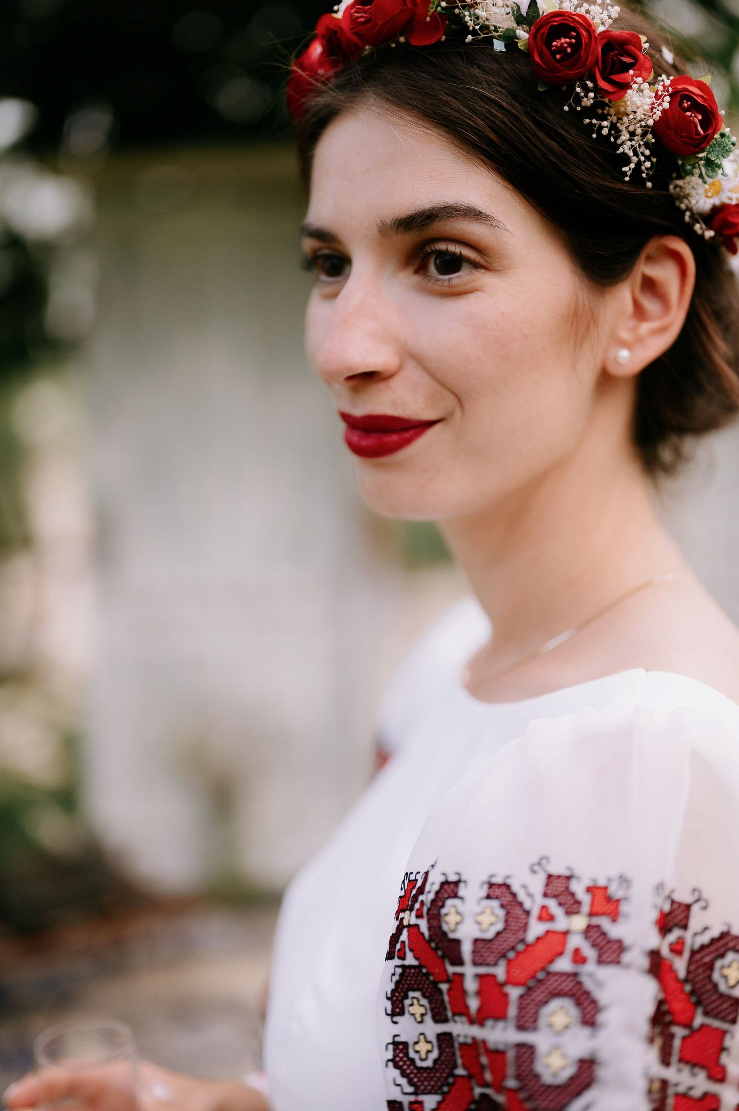 Close-up portrait showing red lipstick and a floral crown with red roses against a blurred background.
