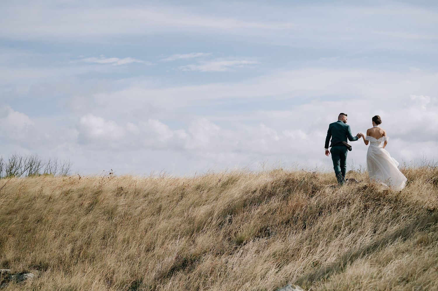 Couple walks along grassy hilltop path during their wedding photoshoot.