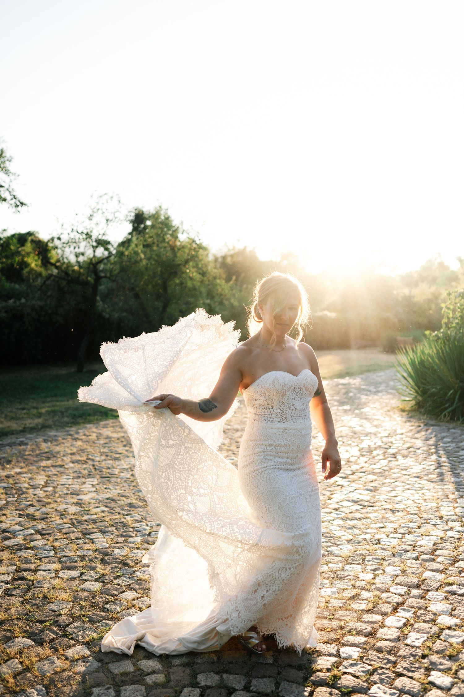 Delicate lace wedding gown billows in the breeze against a sunlit natural backdrop.