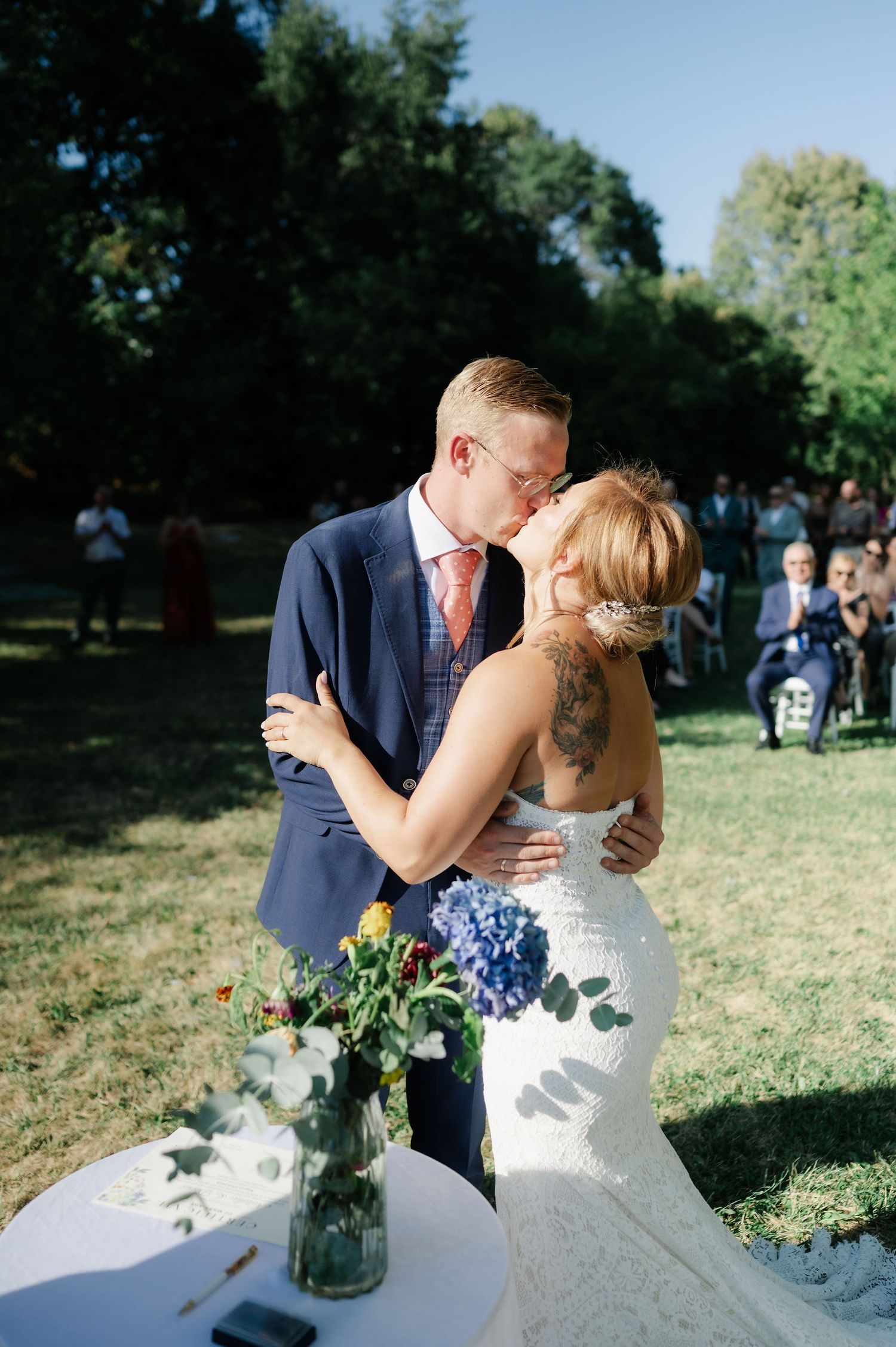 A couple shares a romantic kiss in an outdoor garden setting during a wedding celebration.