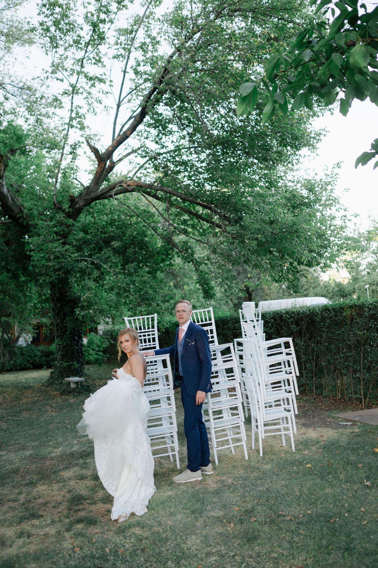 White chairs are arranged outdoors under green trees for a wedding ceremony.