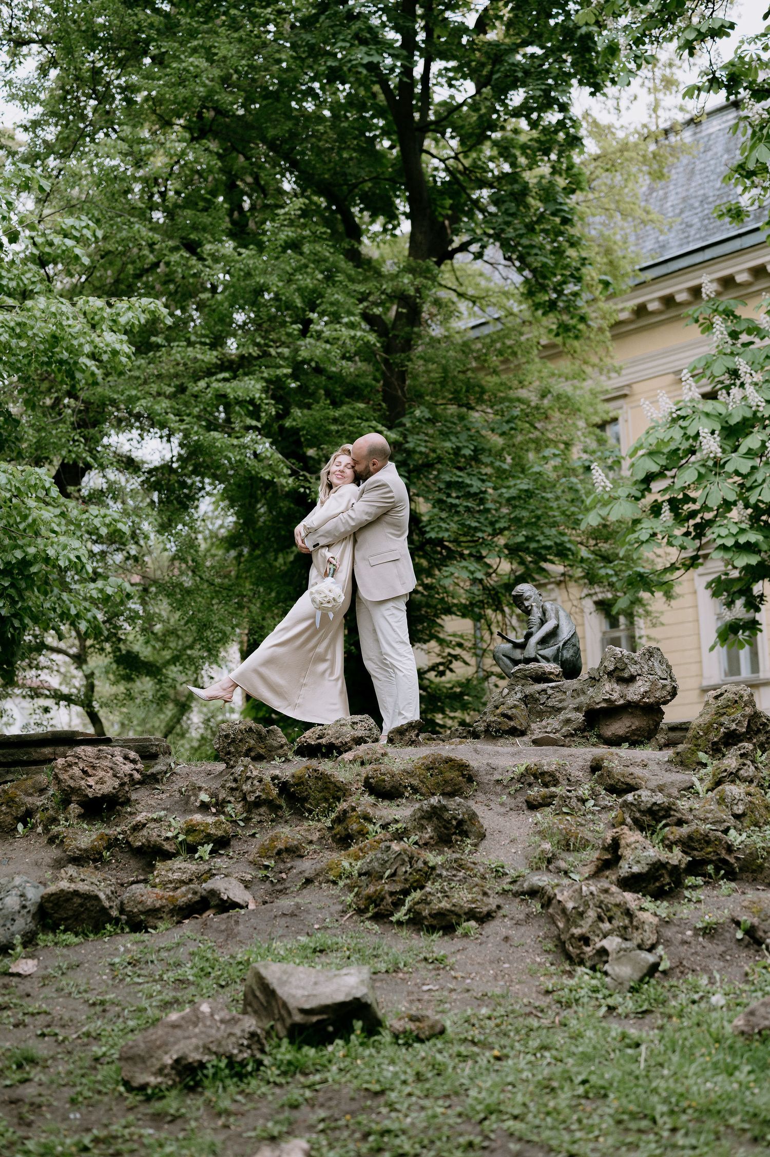 A romantic couple embraces while standing on rocky terrain in a garden setting.