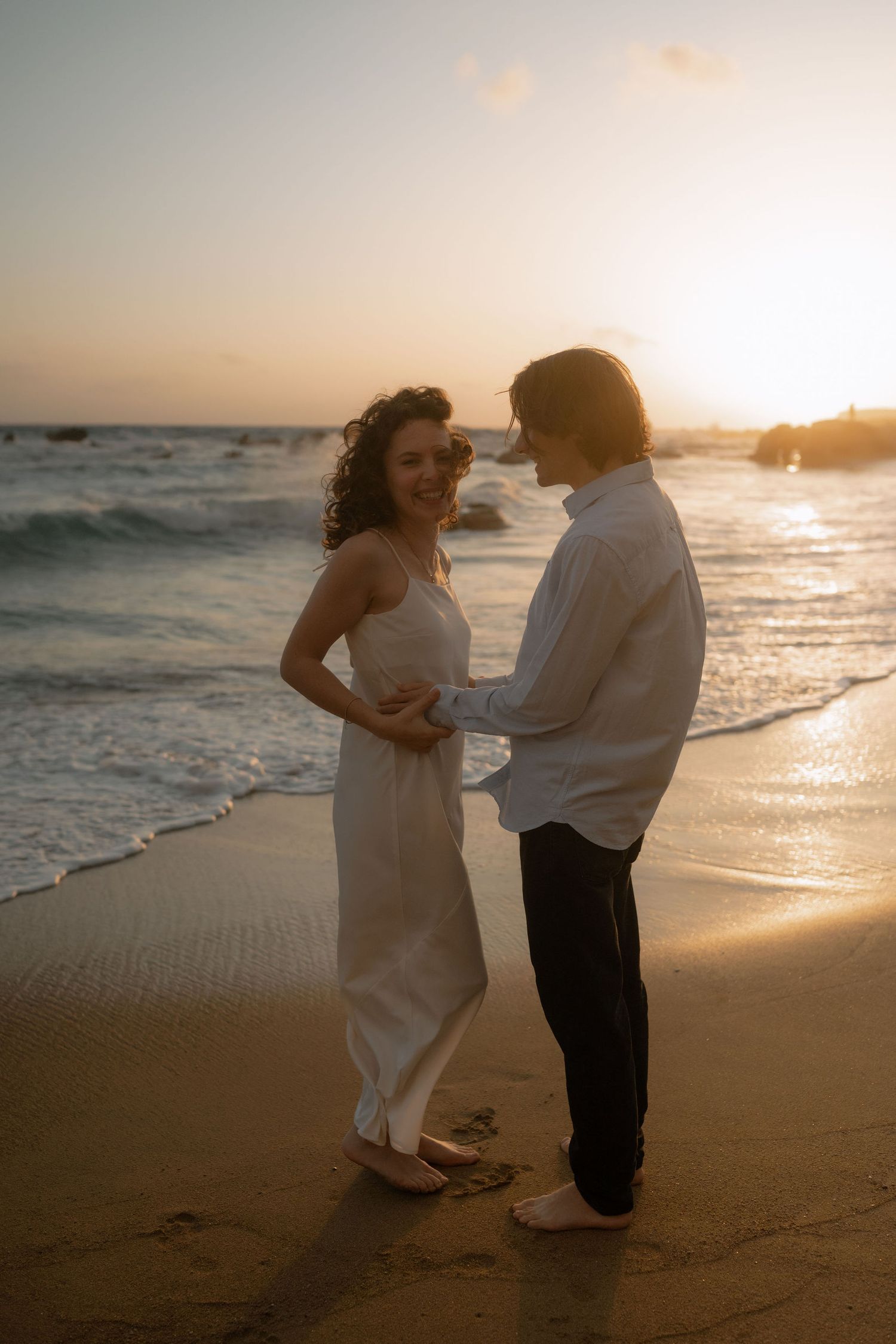 Couple holding hands on a sandy beach at golden sunset, waves gently washing ashore, woman in white dress, romantic moment.