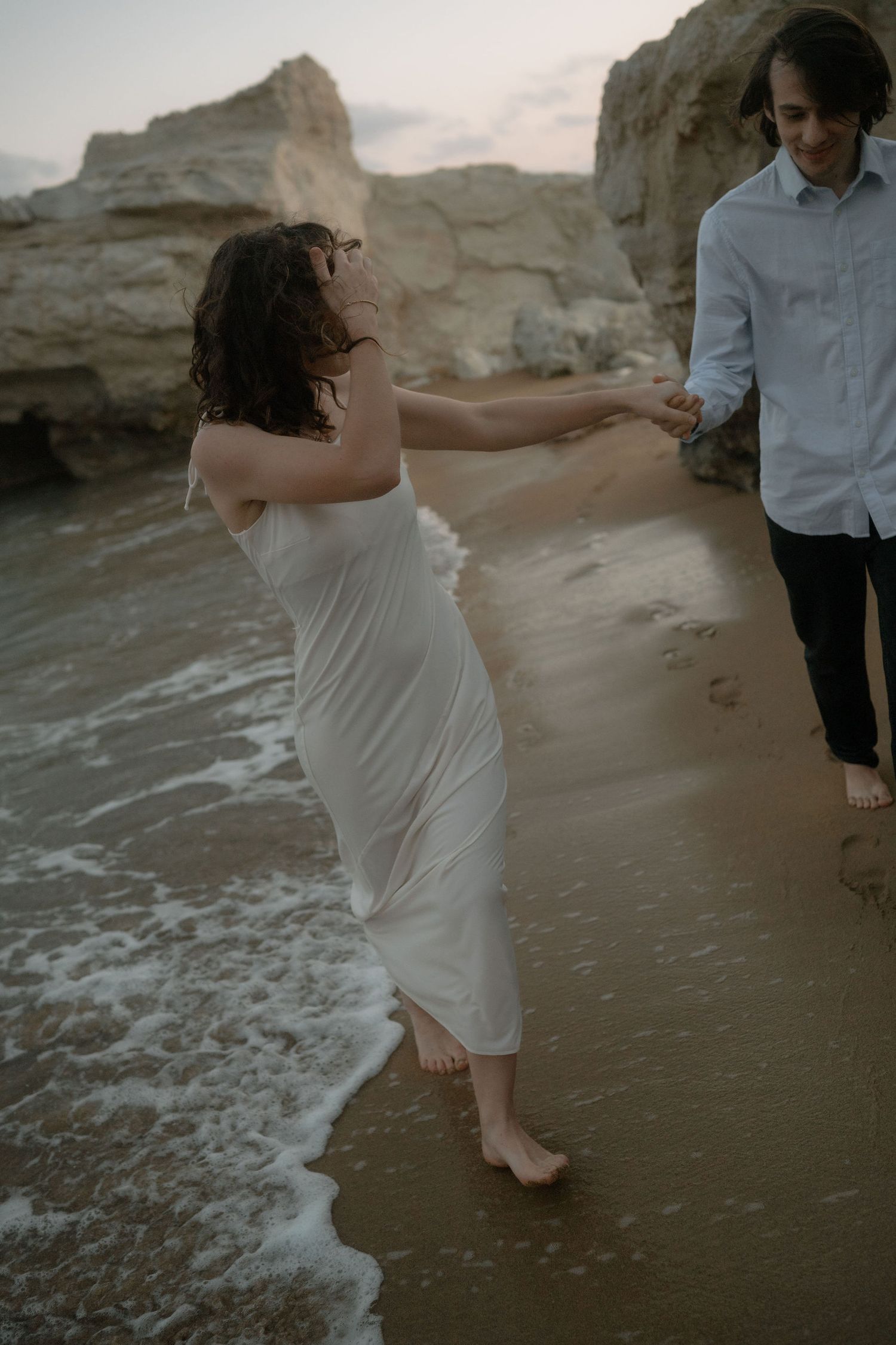 Couple holding hands on a sandy beach at dusk, woman in white dress with ocean waves, rocky cliffs behind them.