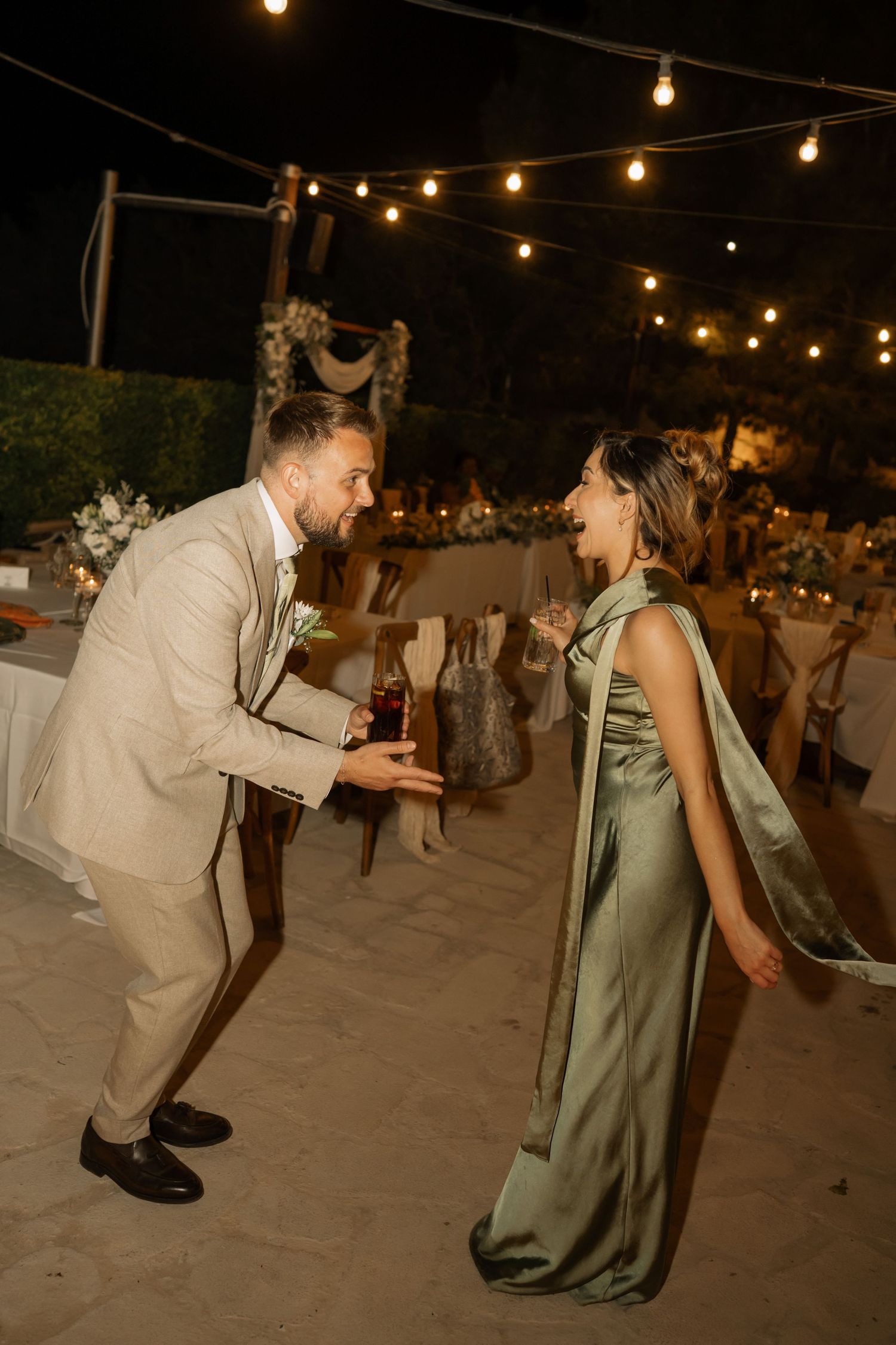 Couple dancing outdoors at night wedding reception under string lights, man in beige suit, woman in green satin gown.