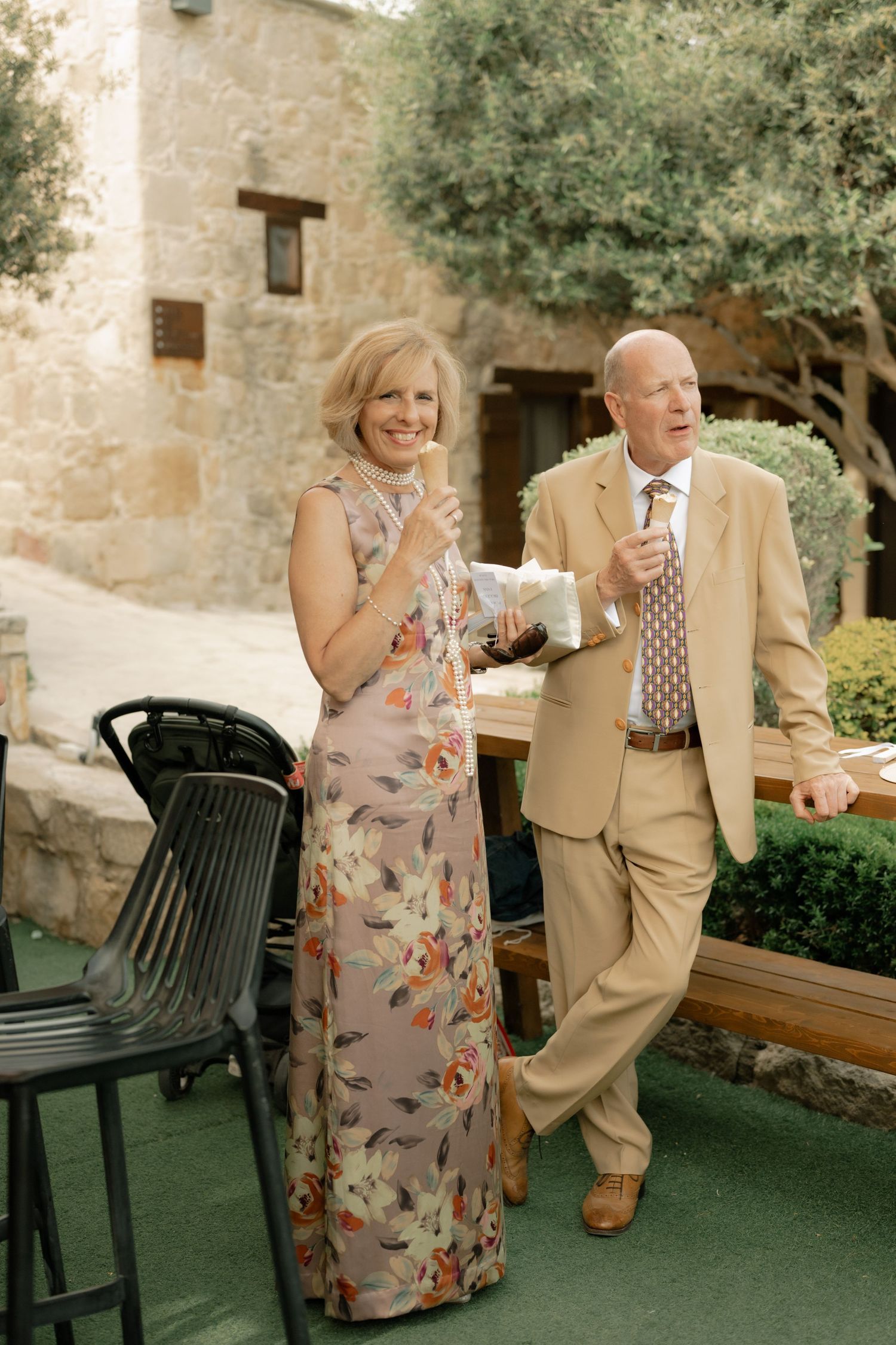 Elegant older couple in formal attire holding drinks outdoors at a stone building venue.