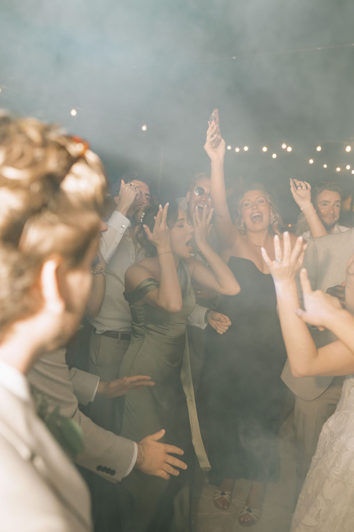 Guests dancing joyfully with hands raised at a wedding reception, surrounded by soft smoke and warm string lights.