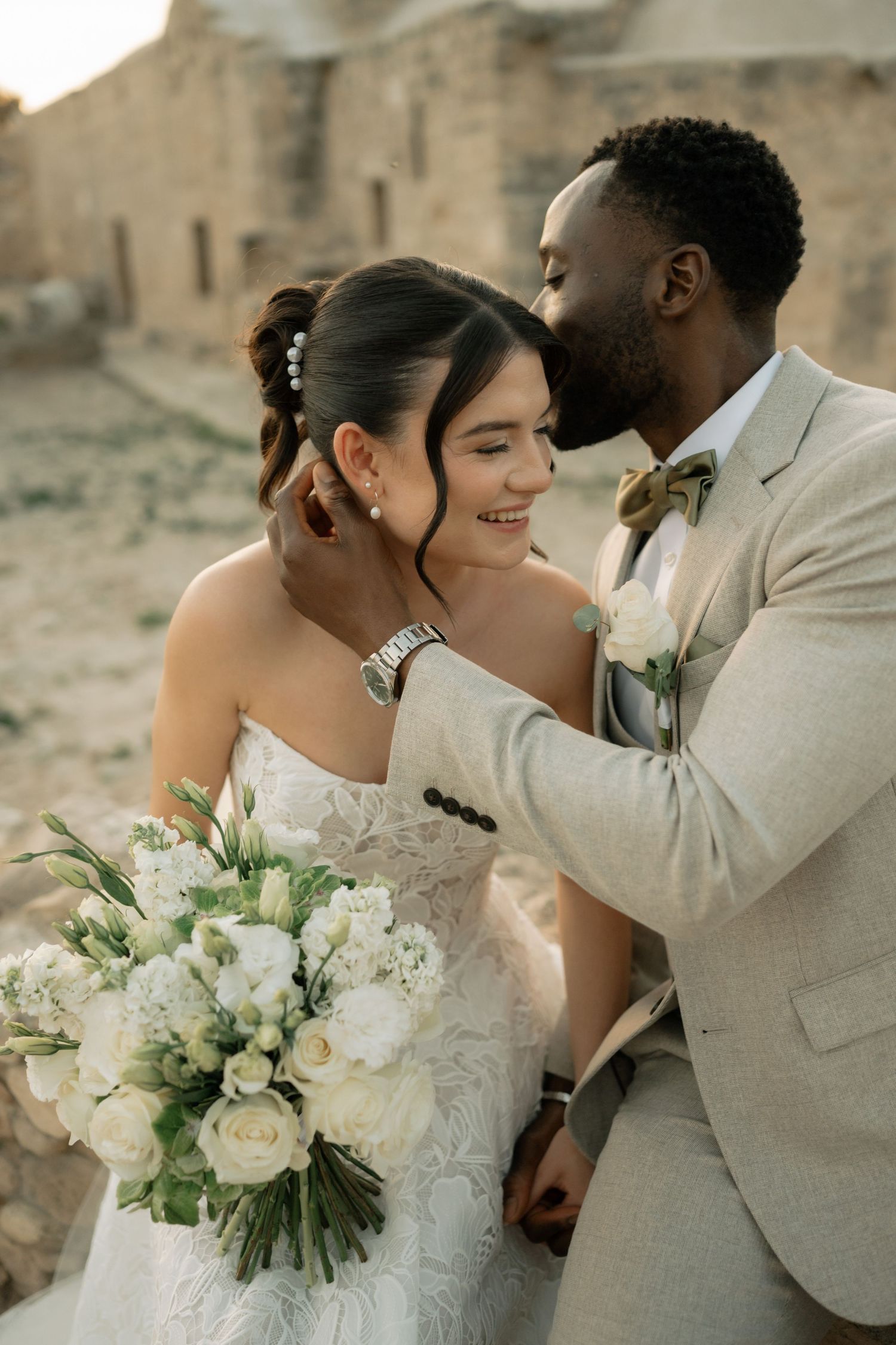 A groom in a beige suit kisses his smiling bride holding white flowers at an outdoor wedding ceremony.