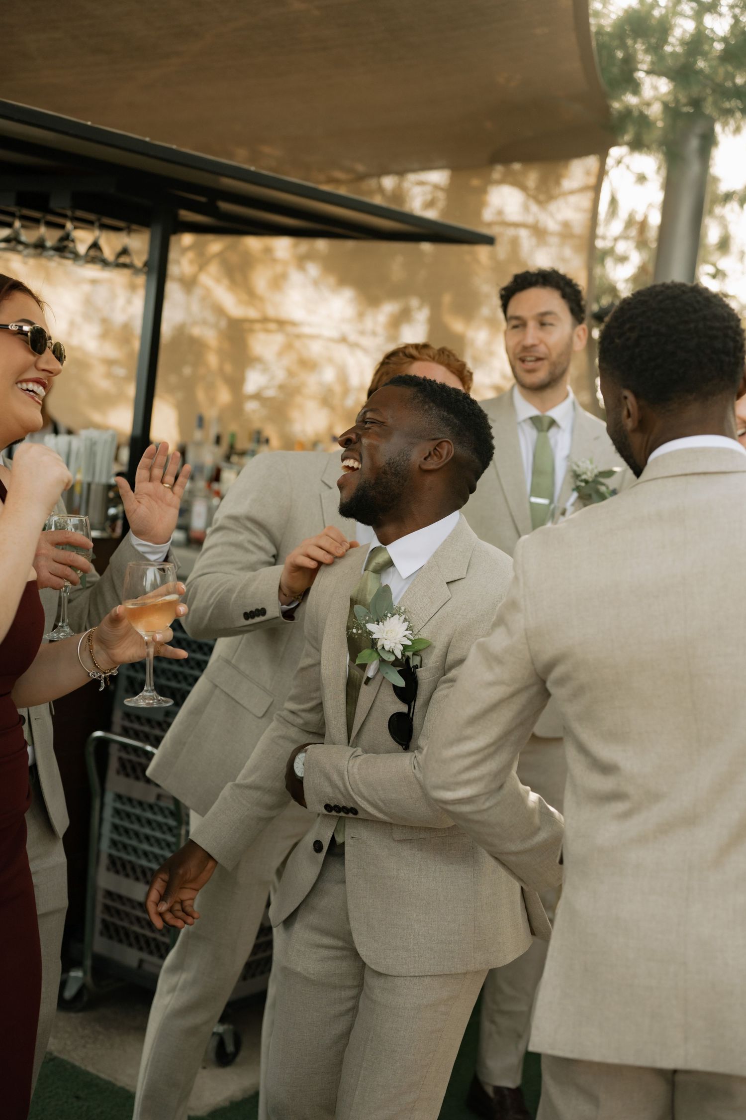 Groom laughing with groomsmen in tan suits at outdoor wedding reception, celebrating with champagne glasses.