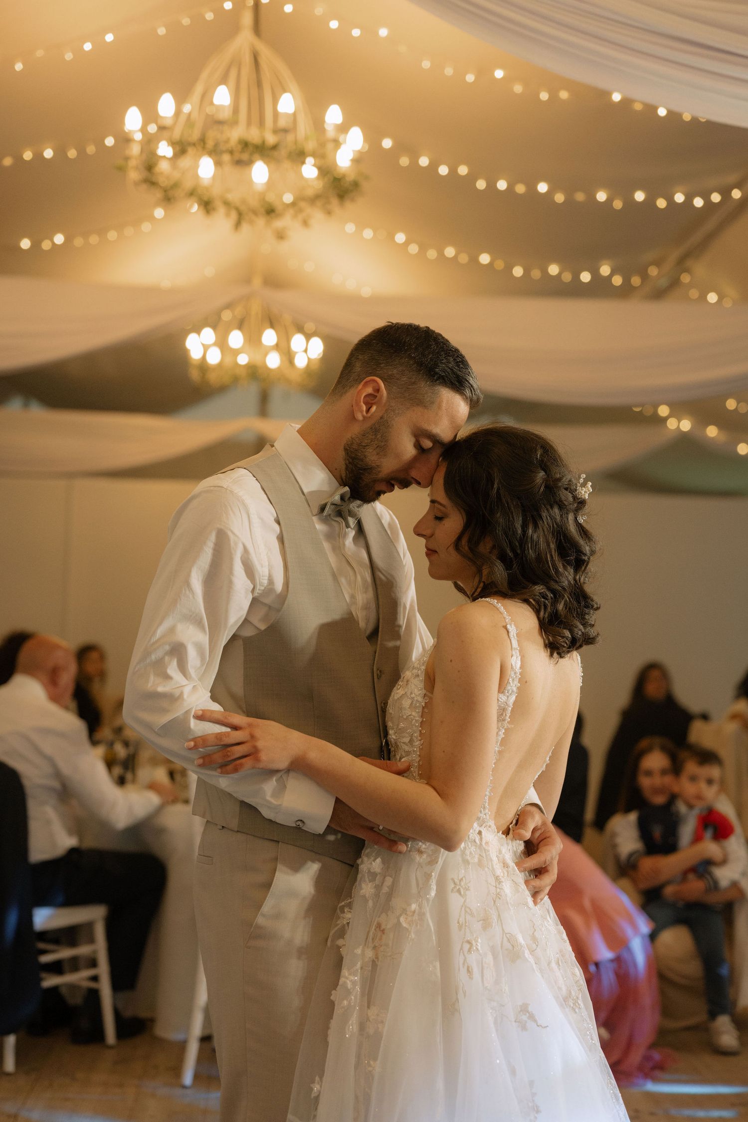 Bride and groom share their first dance under warm chandelier lights and string lights at elegant wedding reception.