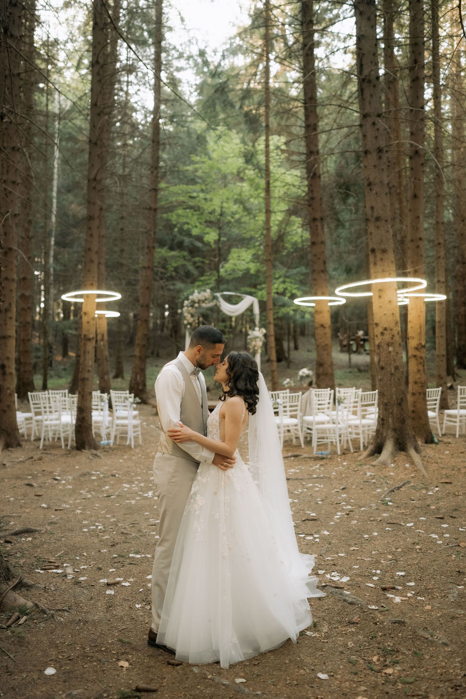 Bride and groom embrace in a enchanting forest wedding venue with glowing ring lights and white chairs.