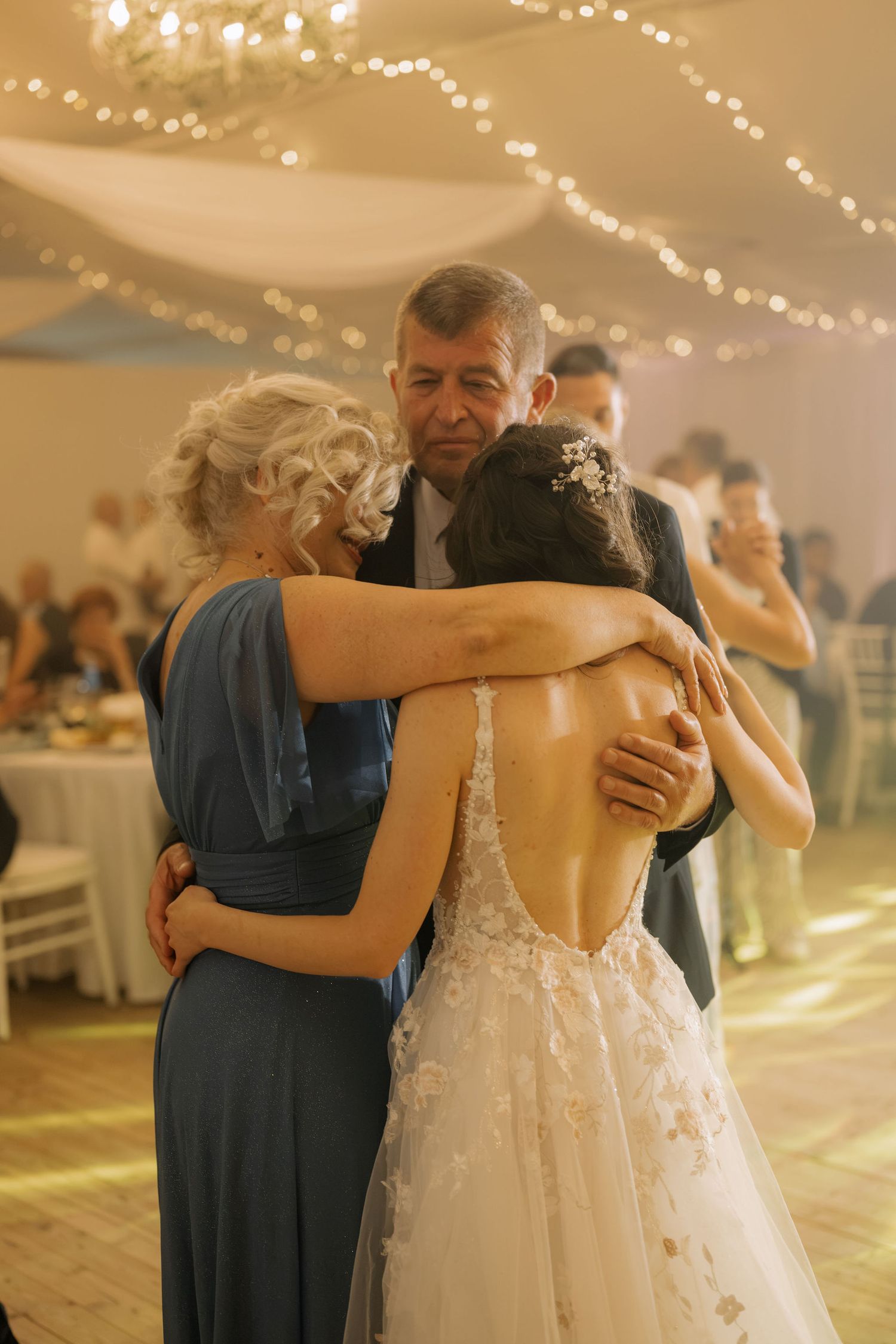 Bride in backless lace gown shares emotional group hug with parents during wedding reception dance.