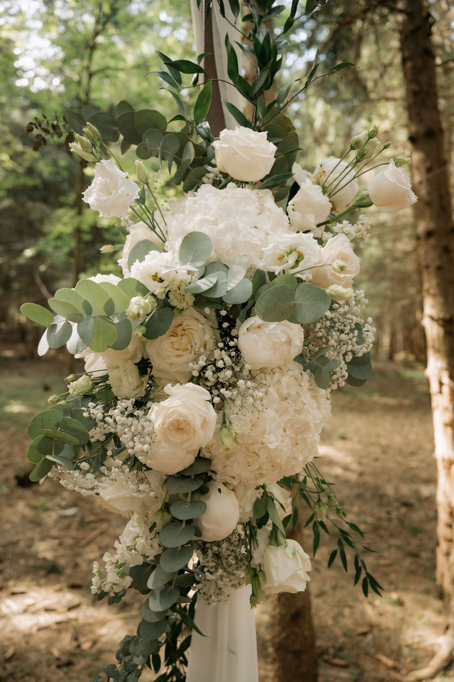 Elegant wedding arch floral arrangement featuring white roses, hydrangeas, and eucalyptus greenery in a lush forest setting.