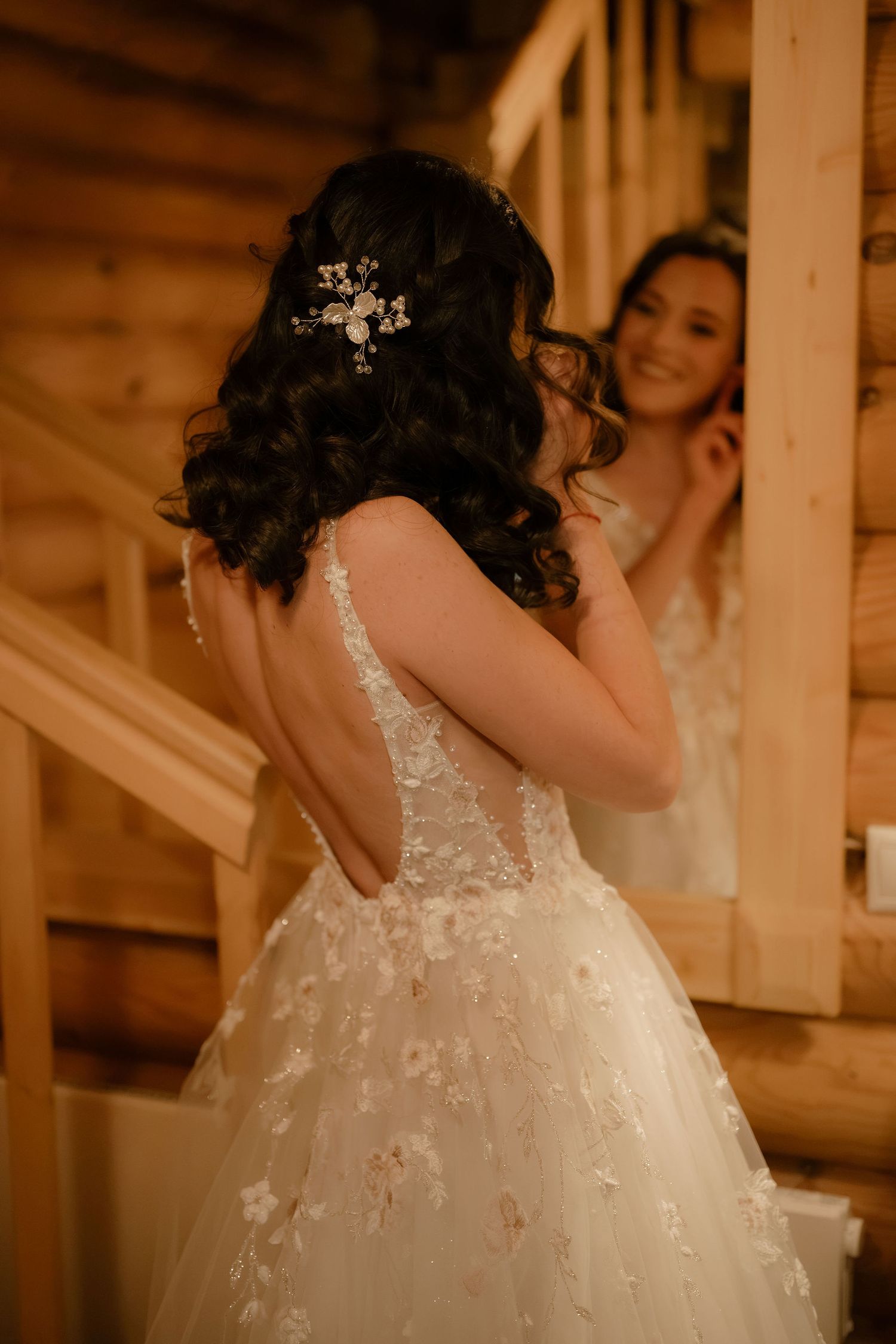 Bride in floral lace wedding gown with hair accessory smiles at her reflection in a rustic wooden cabin mirror.