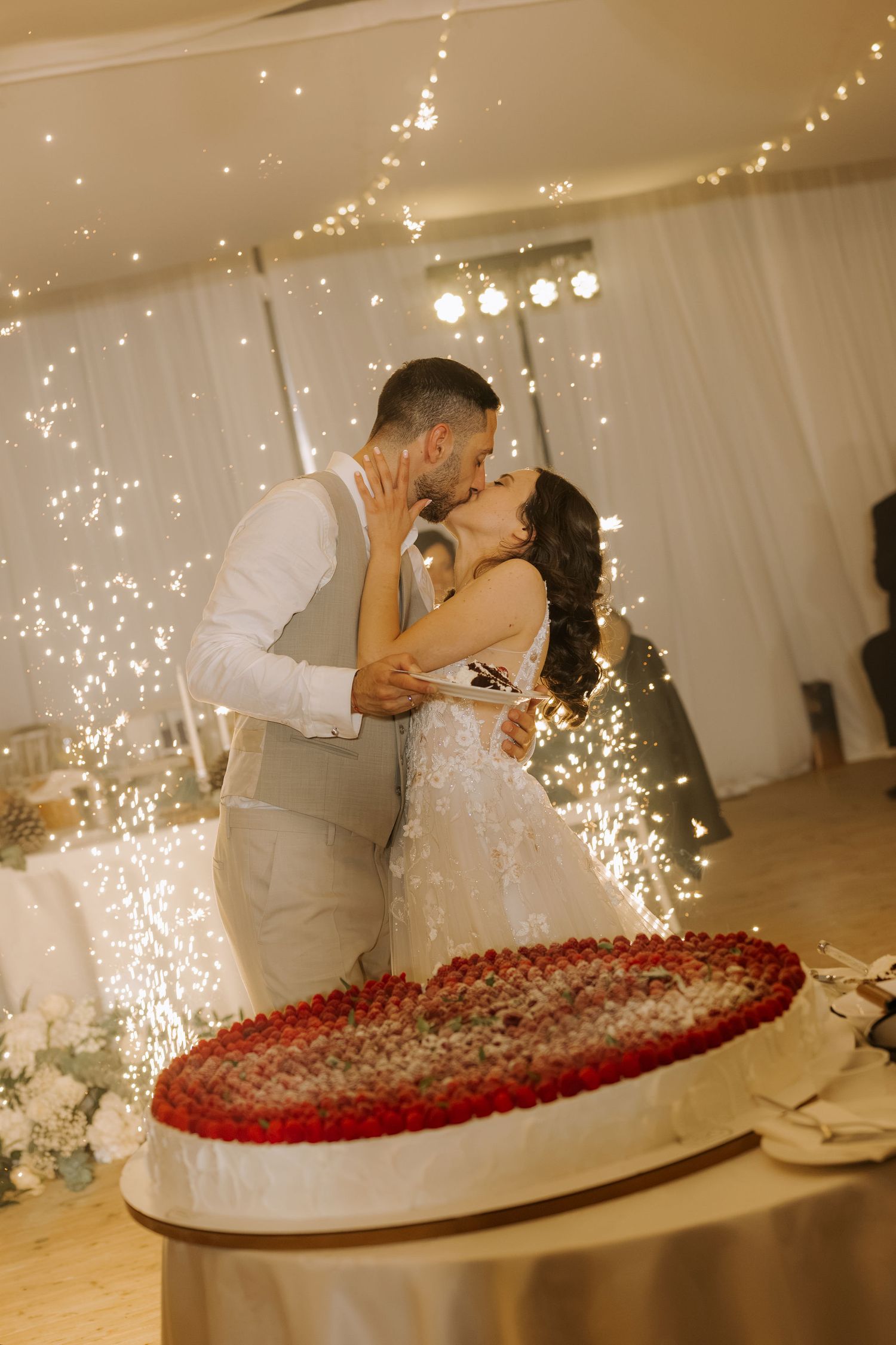 Newlyweds share a romantic kiss in front of a heart-shaped strawberry wedding cake with sparkling fireworks.