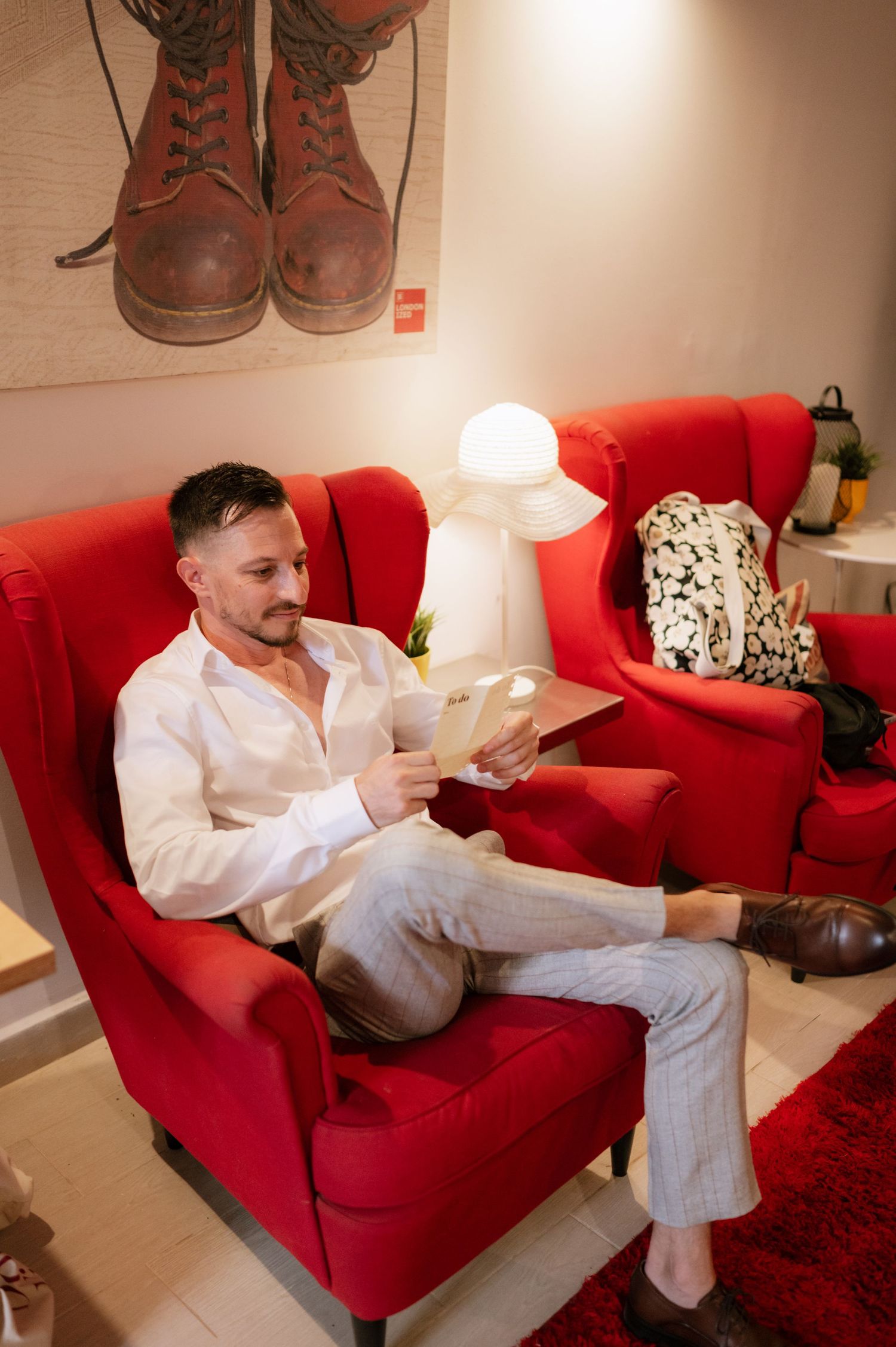 Someone relaxes in a plush red armchair while reading, with a white lamp and decorative boots artwork visible in the background.