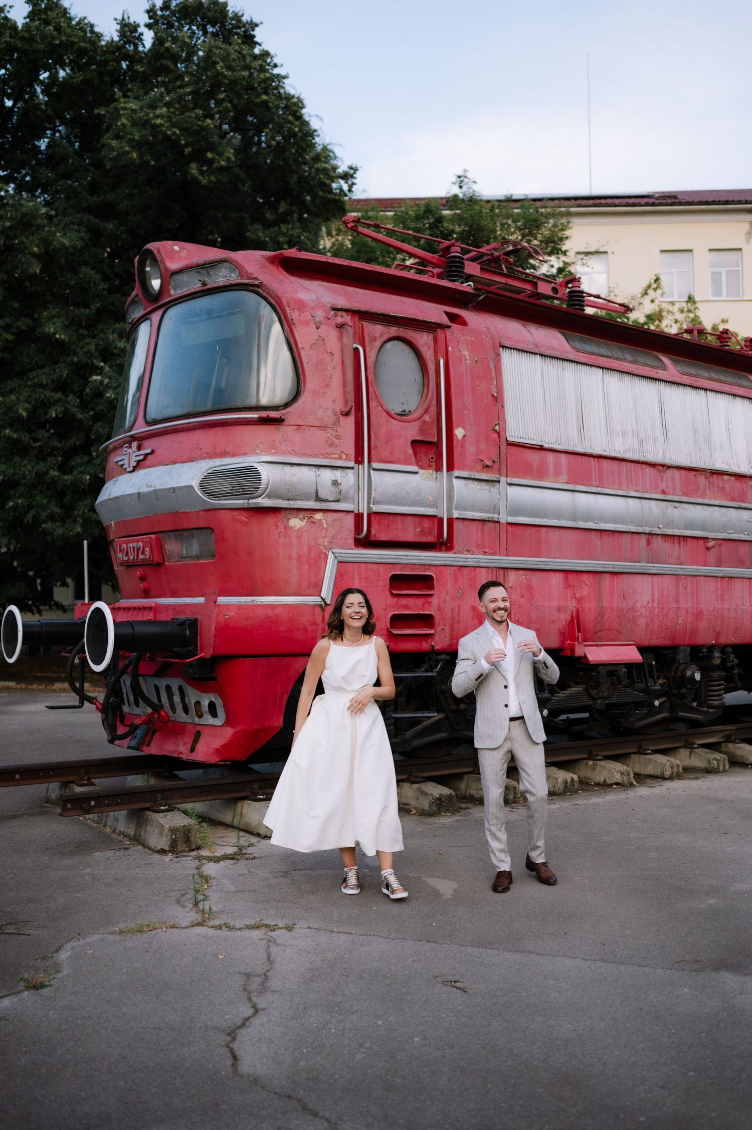 A couple poses in front of a vintage red locomotive at a train museum for wedding photos.