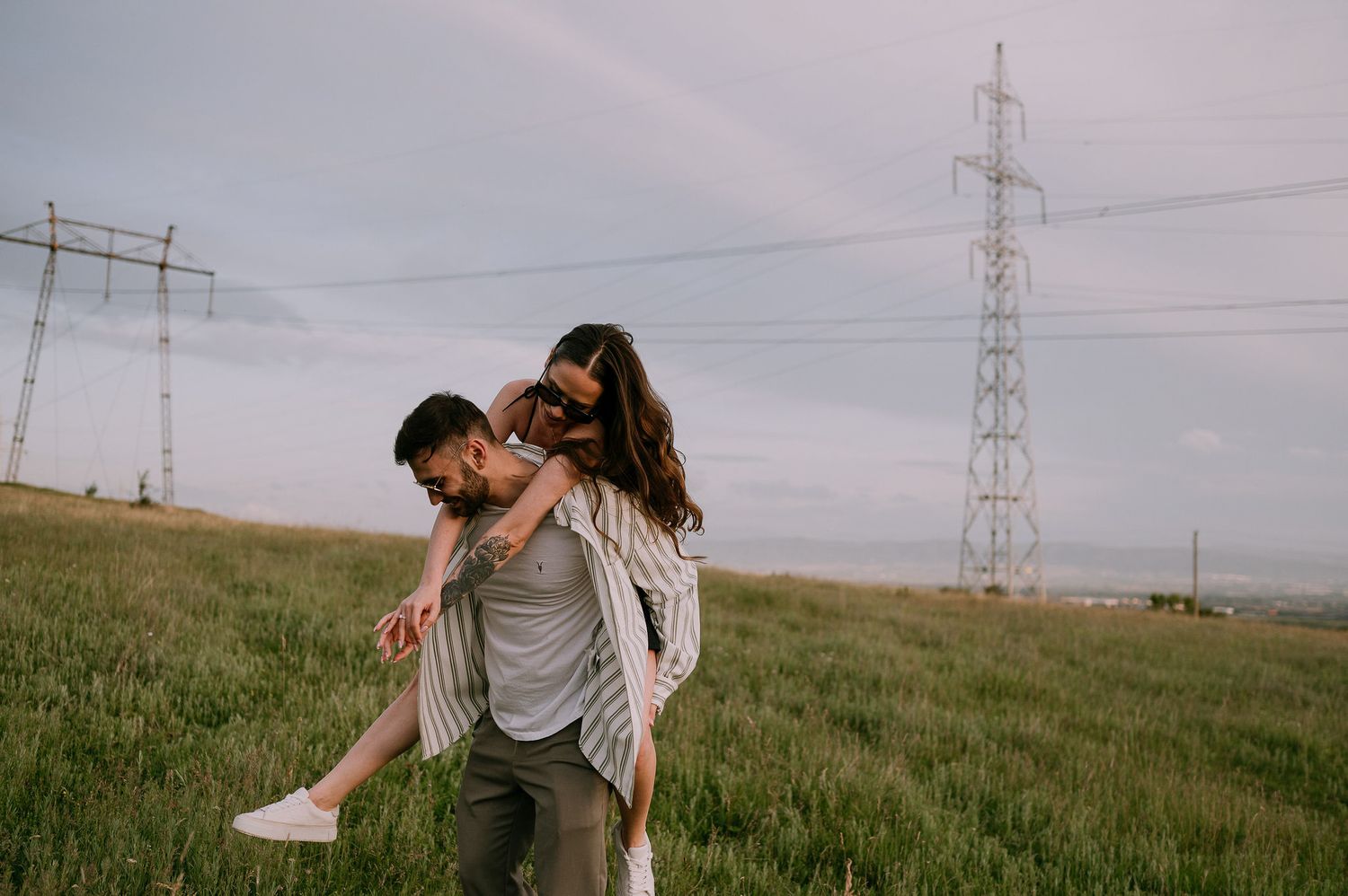 A couple walks together through a grassy field with power lines visible in the background on an overcast day.