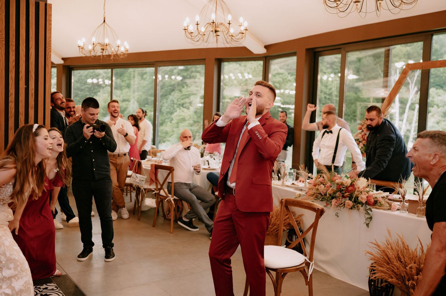 Wedding guests dancing and celebrating in an elegant reception venue with chandeliers and large windows.