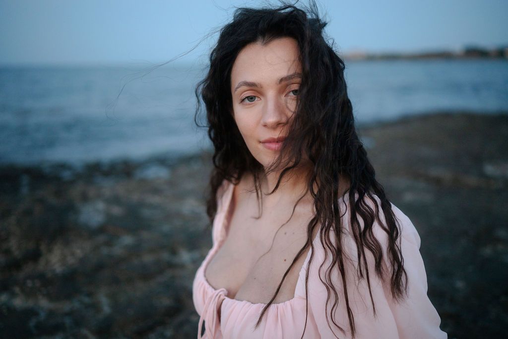 A person with long dark wet hair wearing a pink top stands near the ocean at dusk.