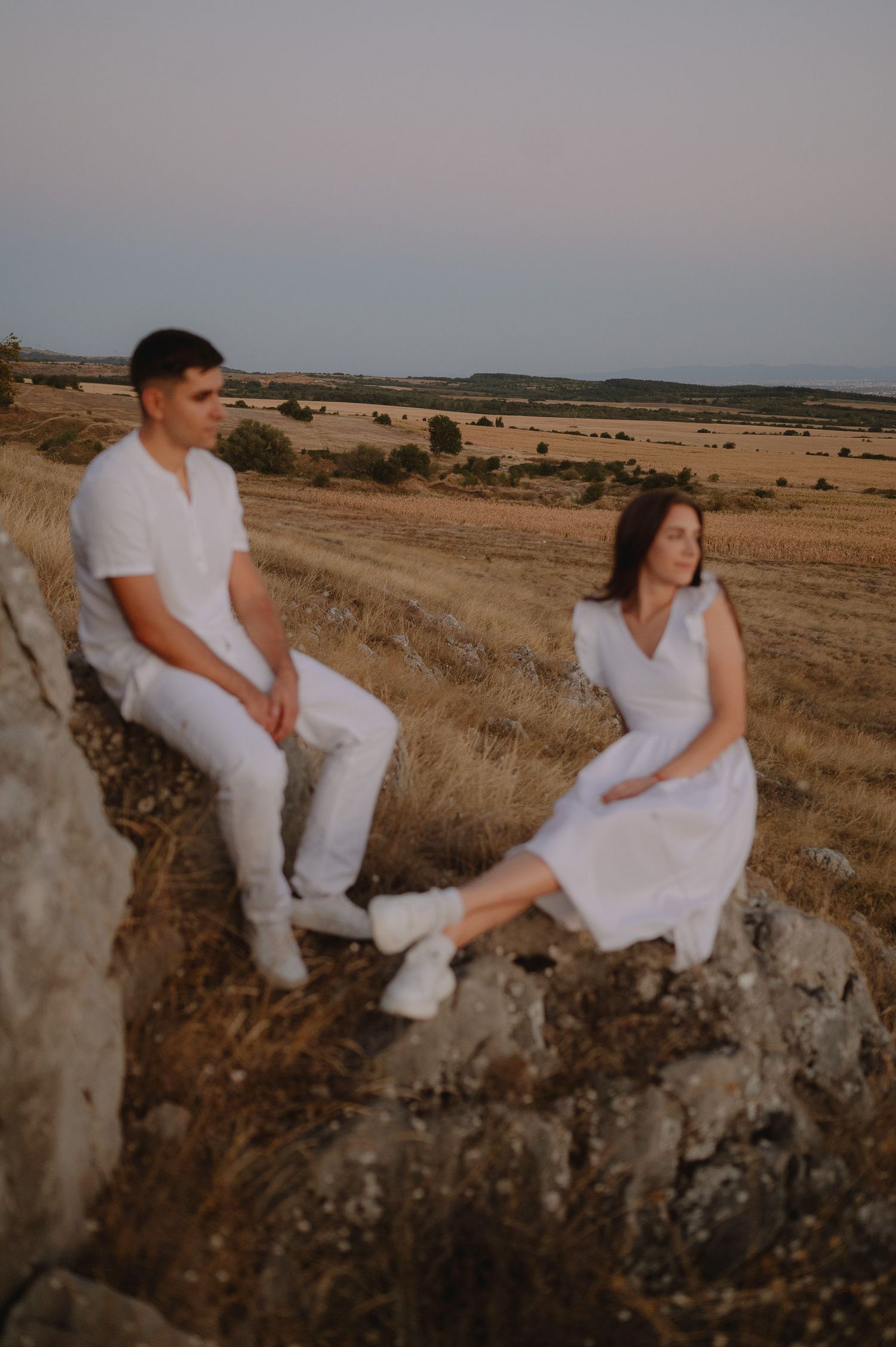 A couple in white outfits sits together on a rocky cliff edge overlooking a vast rural landscape at sunset.