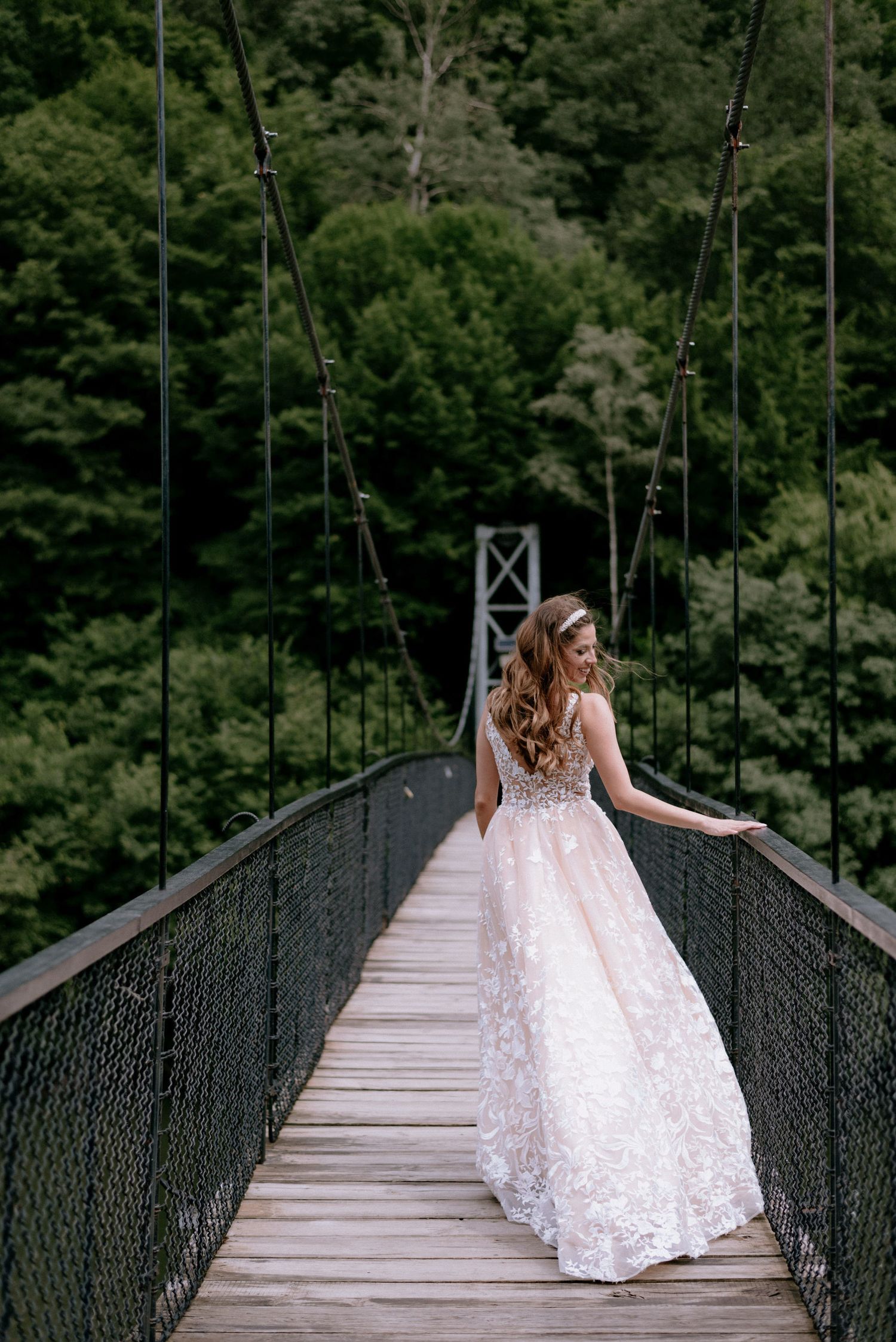 A bride in an elegant white lace gown stands on a rustic suspension bridge surrounded by lush green forest.