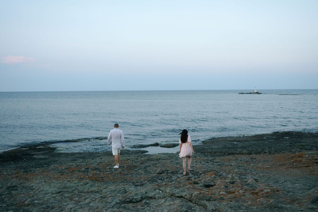 Panoramic seascape showing two figures walking along a rocky shoreline at dusk with calm ocean waters stretching to horizon.