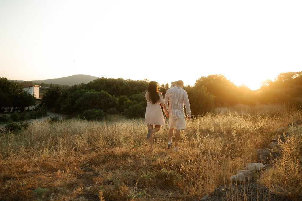 Two people holding hands walk through a golden field at sunset with mountains in the background.