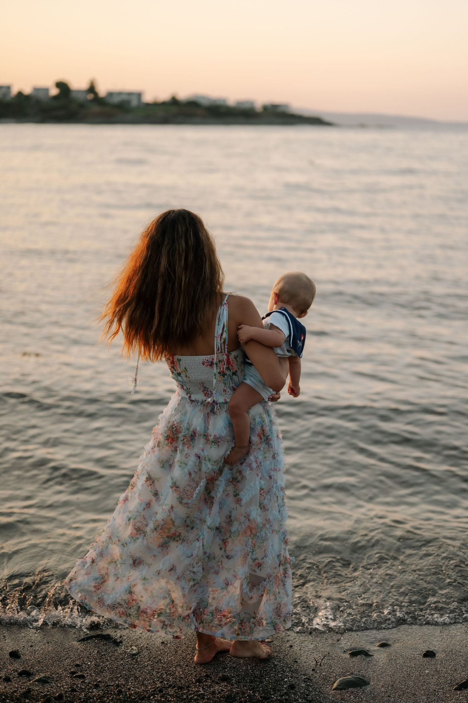 A figure in a floral dress sits on a rocky beach at sunset with waves lapping at the shore.