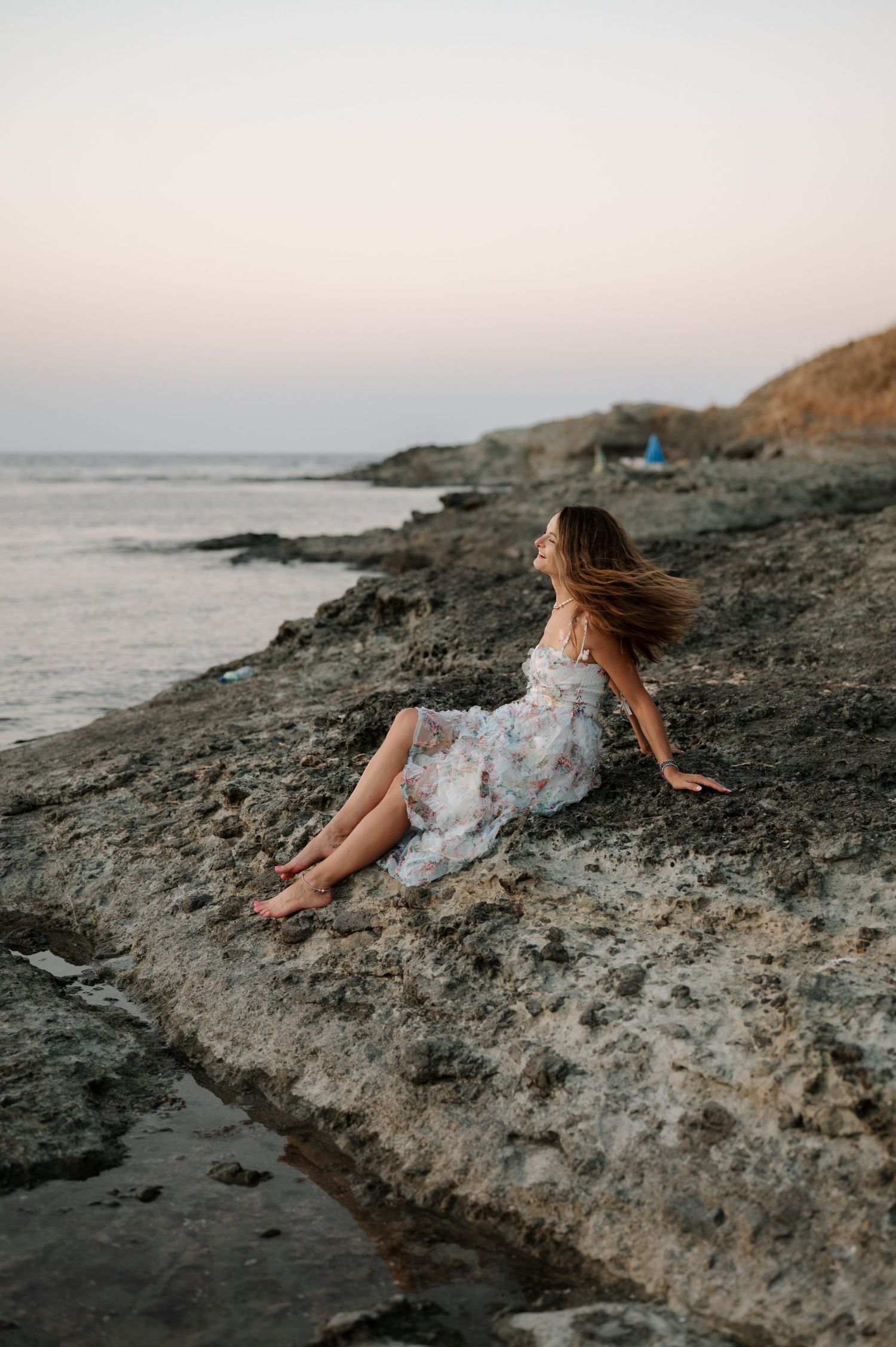 Someone in a white dress sits on rocky coastal cliffs at sunset as their hair blows in the wind by the ocean.