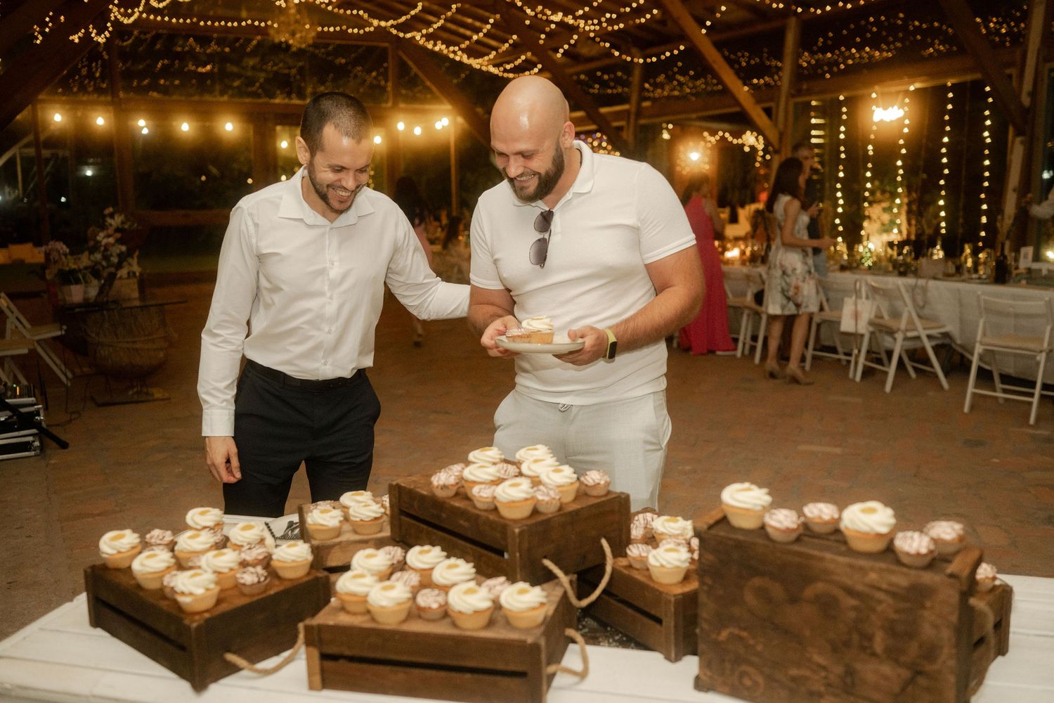 People cut their wedding cake together while guests watch during an evening reception with warm lighting and decorations.