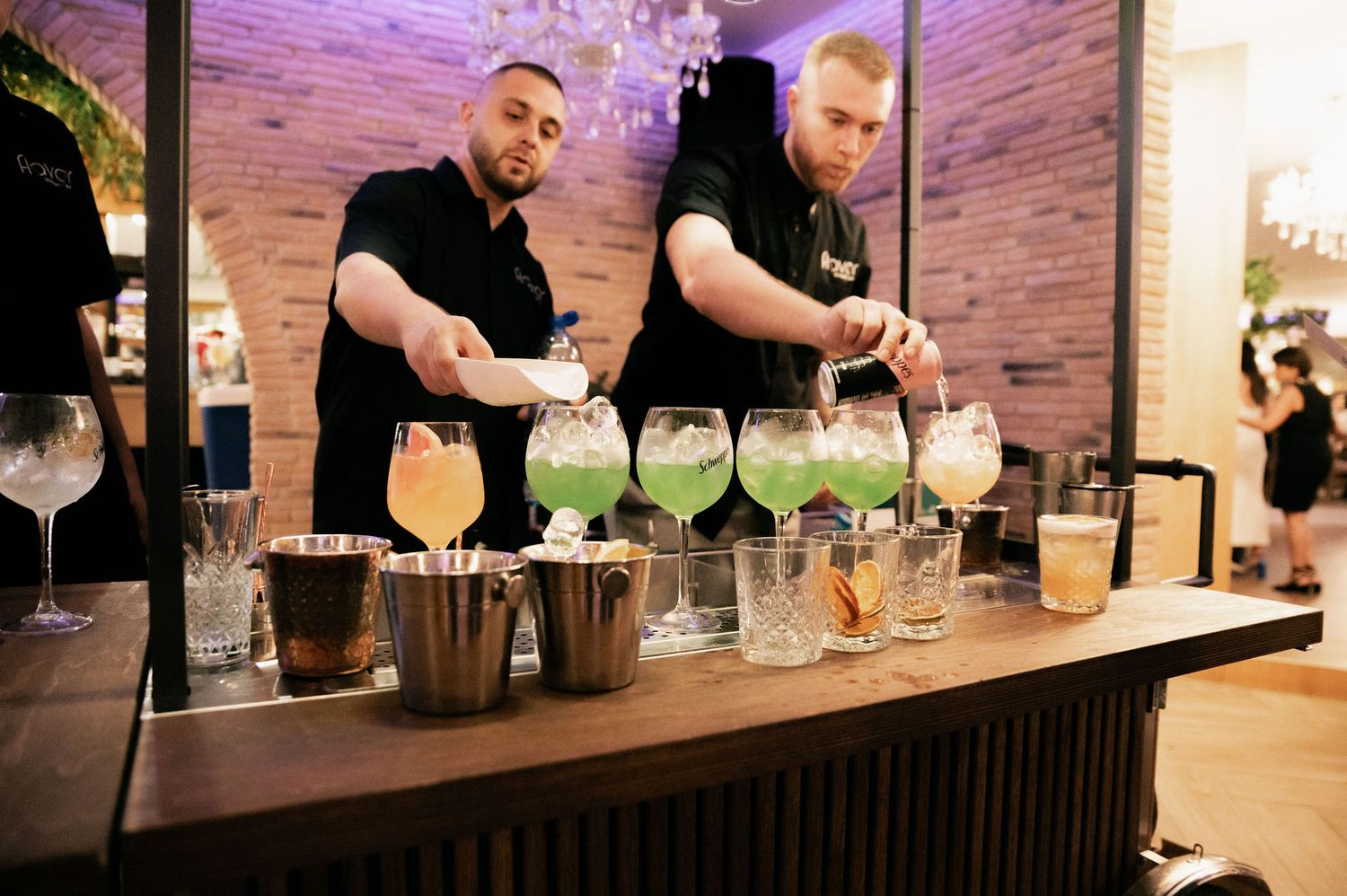 Bartenders prepare cocktails behind a wooden bar counter with glass containers and purple lighting in the background.