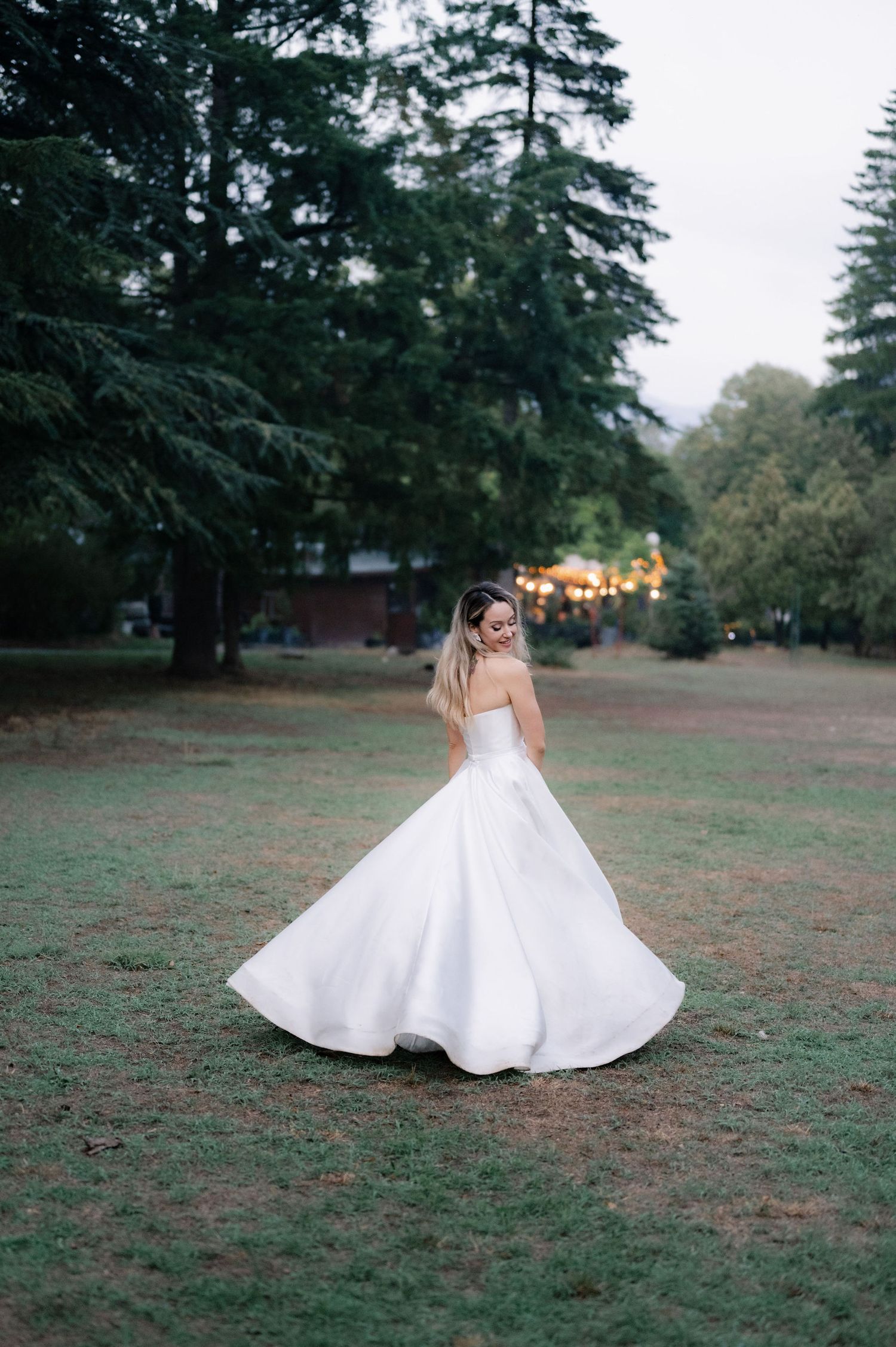 A white bridal gown spins elegantly on a grassy lawn with evergreen trees creating a natural backdrop.