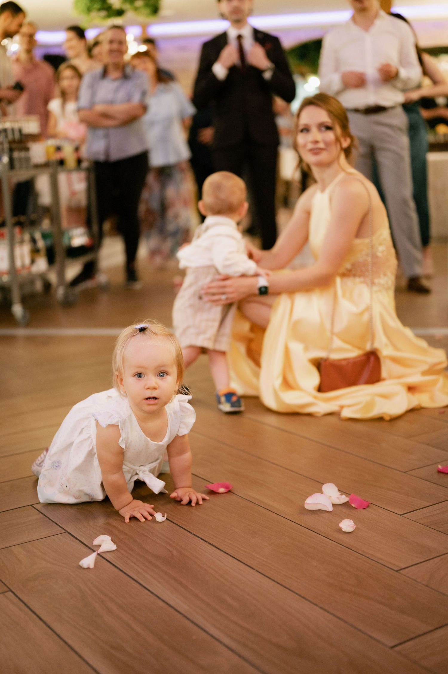 A toddler in white dress crawls on wooden dance floor while flower petals are scattered during a wedding reception.