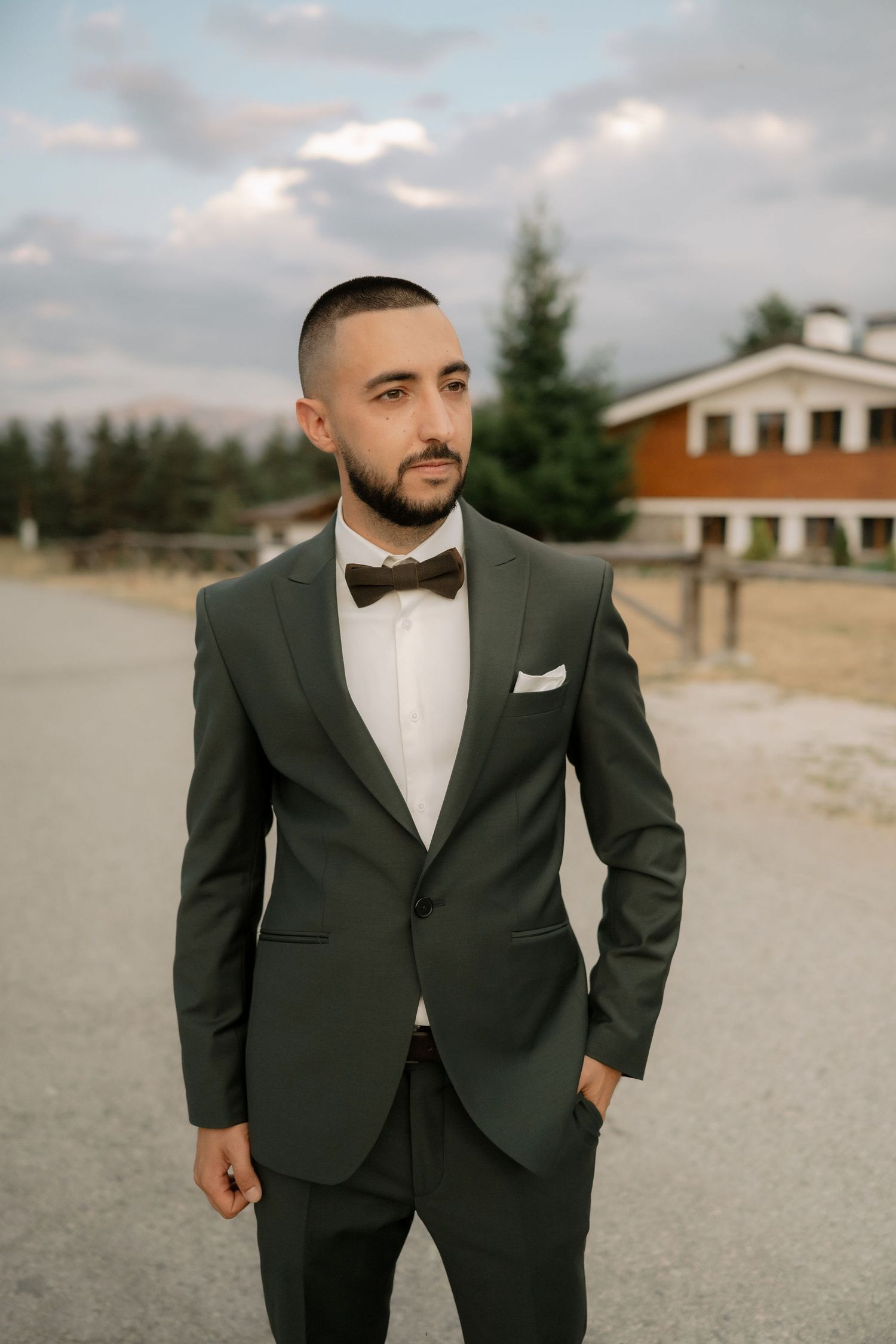 A person in a classic black tuxedo and bow tie poses outdoors on a cloudy day.