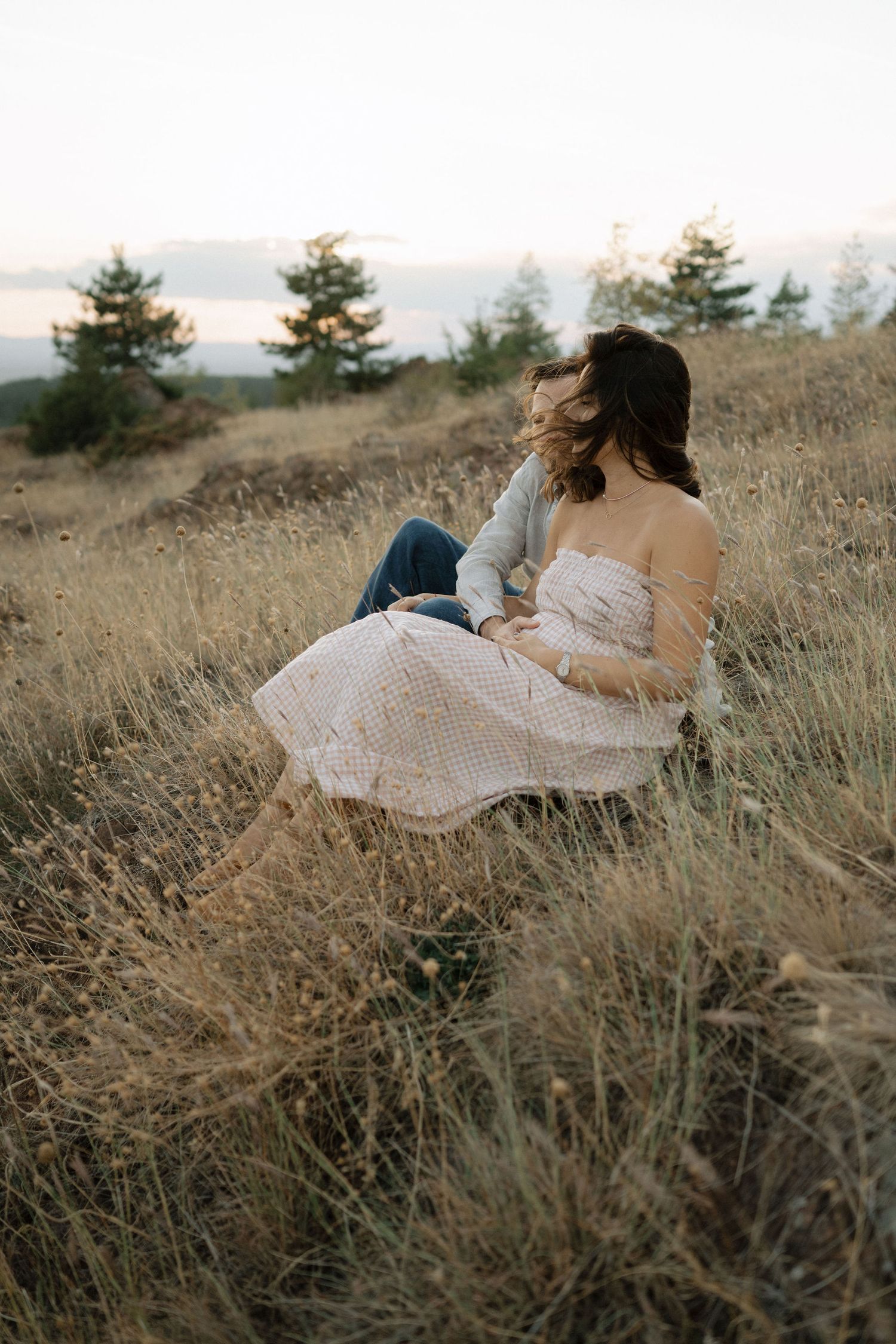 A couple sits together in a golden field of tall grass during sunset while wearing romantic white attire.