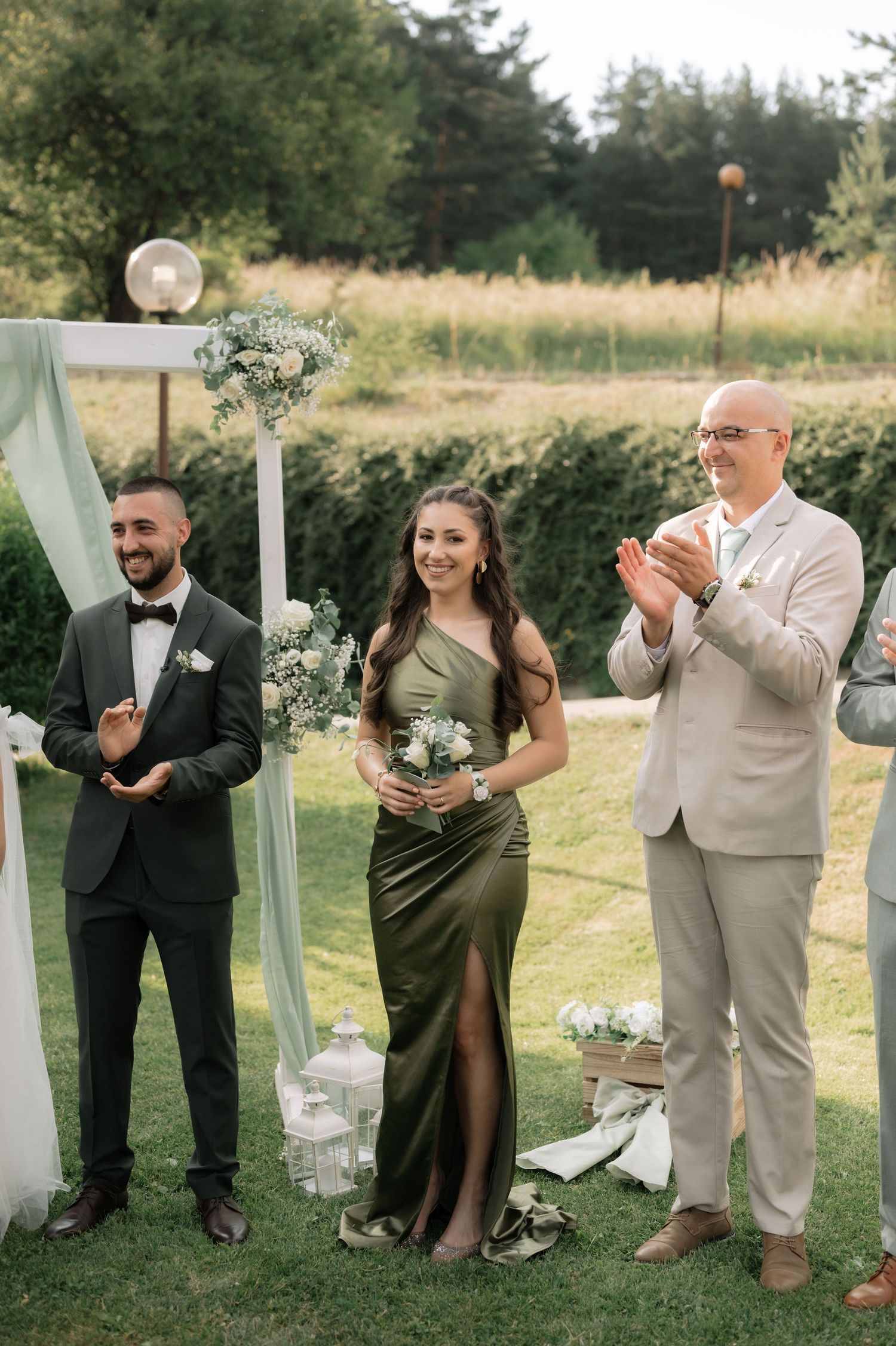 Wedding party members in formal attire pose together on a lawn with sage green and white decor.