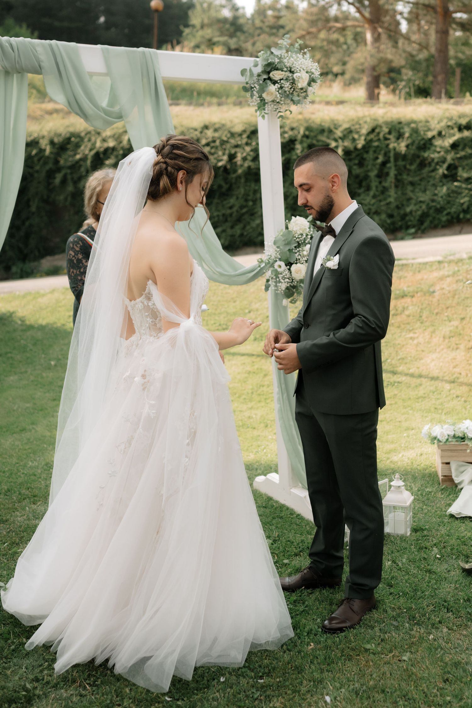 A wedding ceremony takes place outdoors with a flowy mint green arch and white lanterns on a grassy lawn.