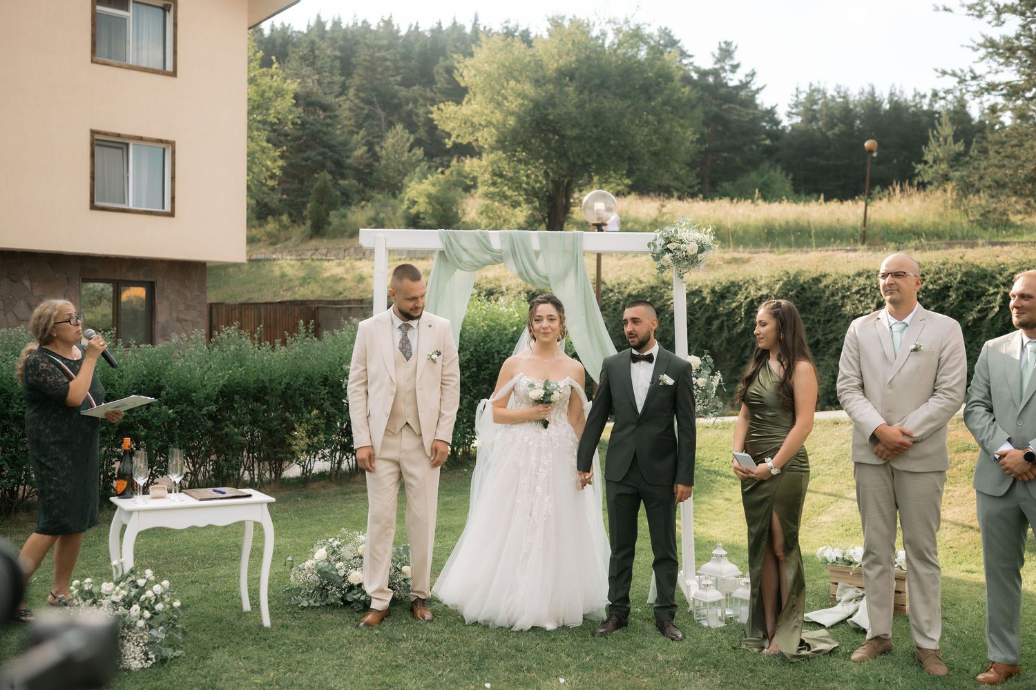 An outdoor wedding ceremony setup with a small wedding party gathered on a lawn near evergreen trees and a building.