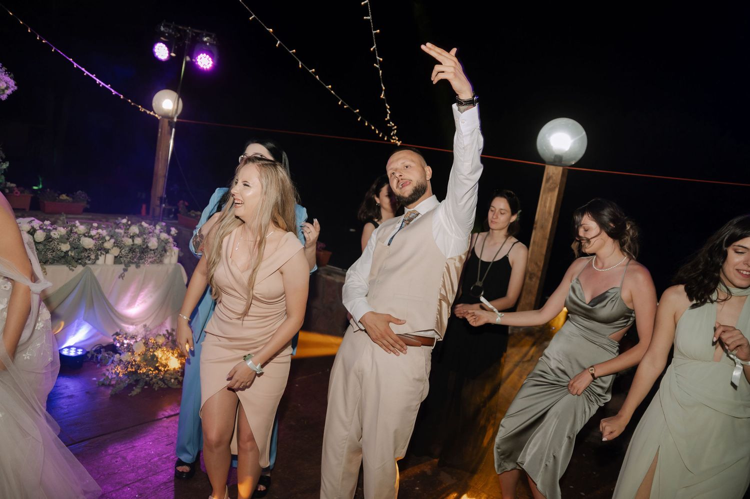 Wedding guests dance and celebrate on a dimly lit dance floor with purple lighting and decorative elements.