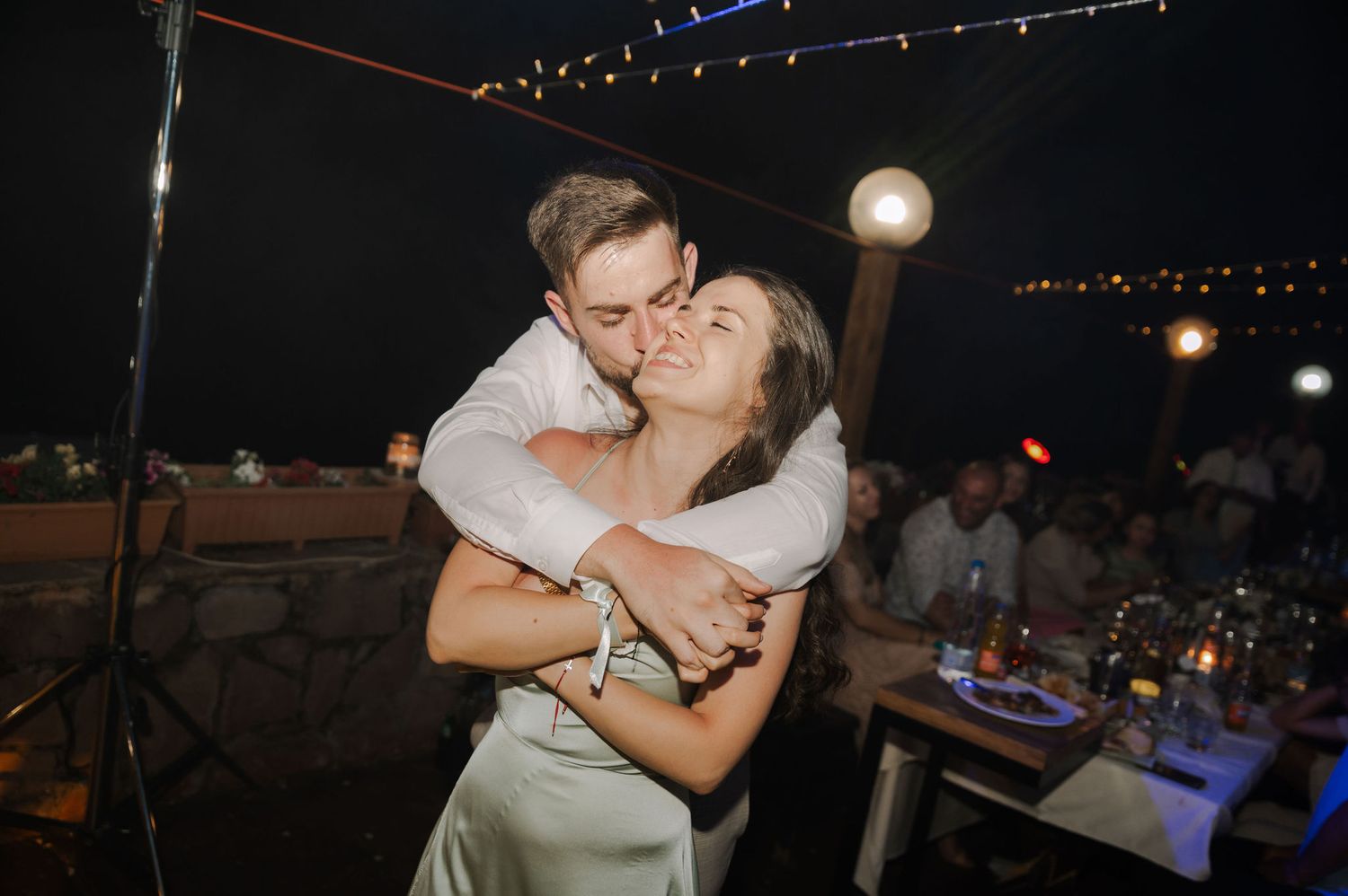 Couple embraces and kisses during a romantic nighttime celebration under string lights at an outdoor venue.