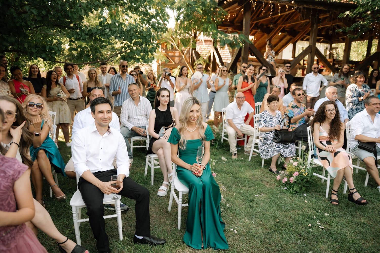 Wedding guests seated outdoors on white chairs during a garden ceremony with green trees in the background.