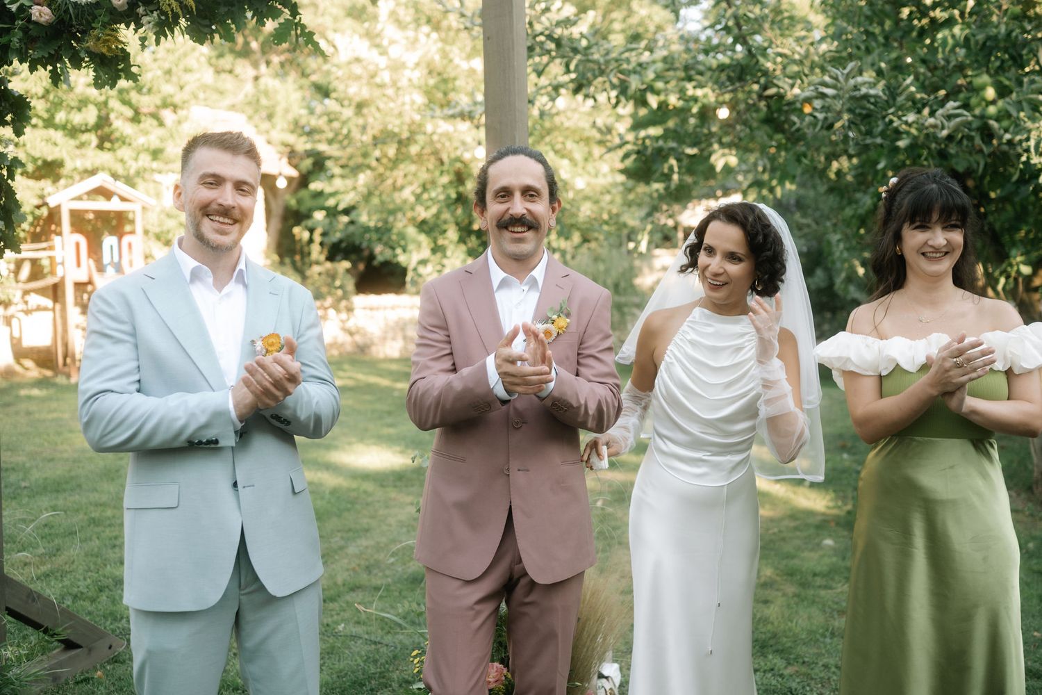 Wedding party members clap and celebrate together in a sunny outdoor garden setting.