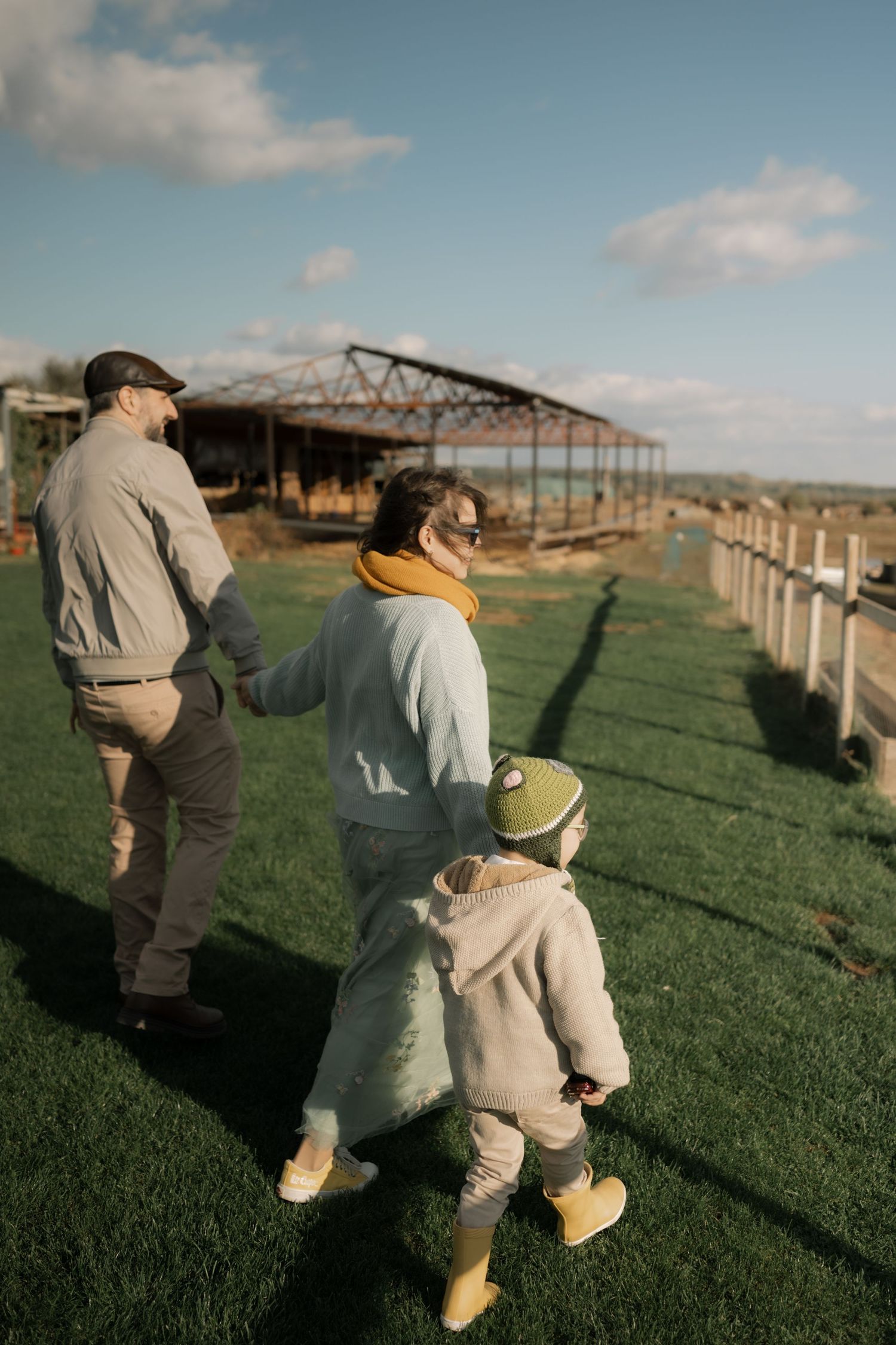 Two people walk along a grassy field next to a metal structure under a partly cloudy sky.