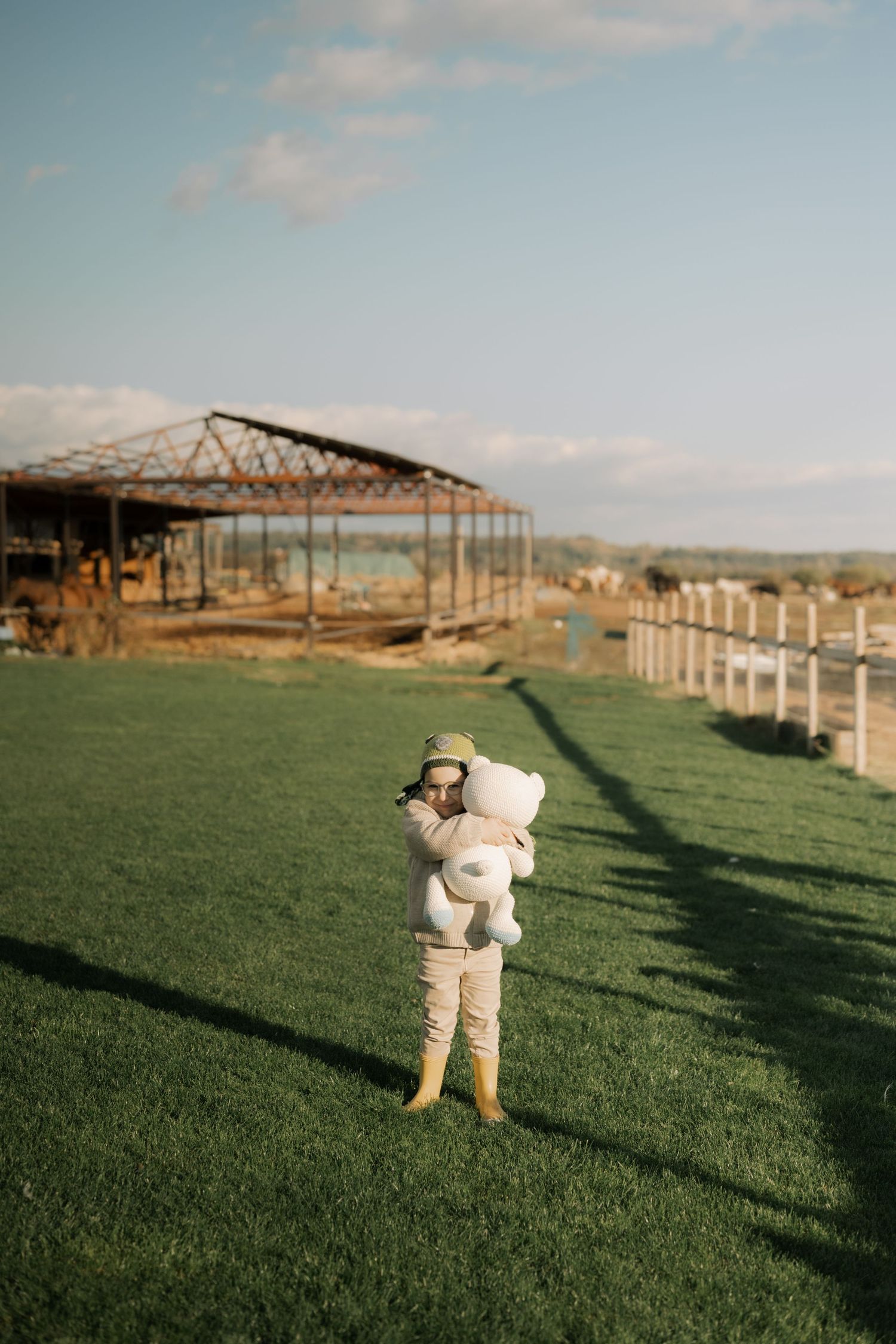 A person in a white outfit and hat stands in a green field next to an old wooden structure on a sunny day.