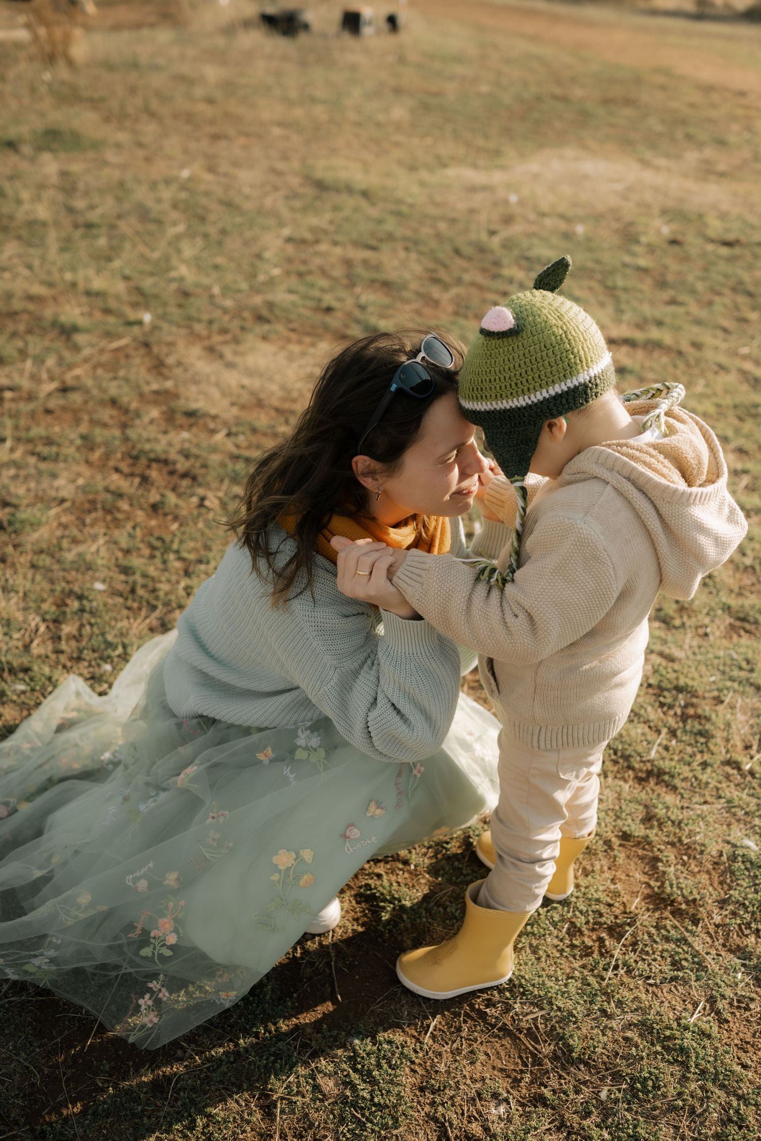 A tender moment captured outdoors as a child in yellow boots and a knit hat interacts with someone in a flowy dress.